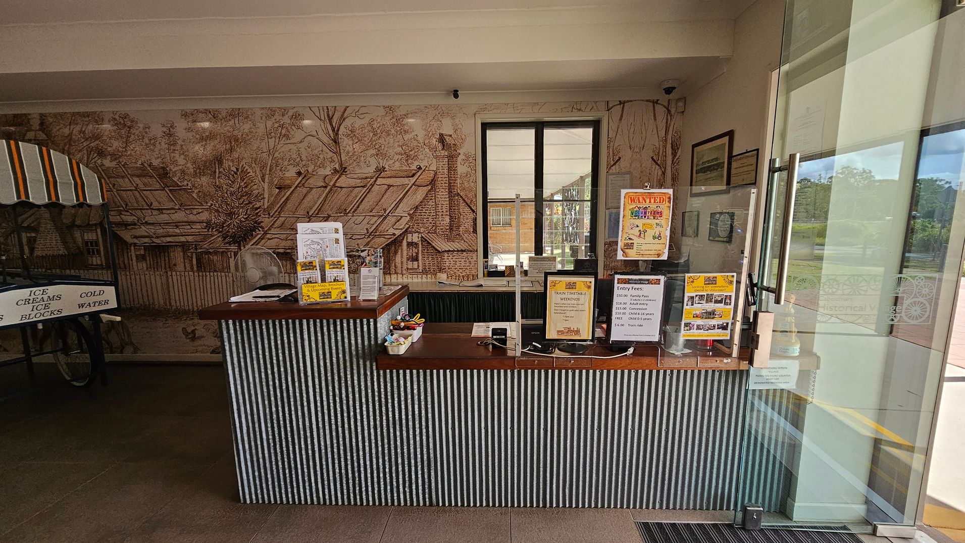 A reception area with a corrugated metal counter displaying various brochures and signs, including entry fees and a volunteer notice. Behind the counter, a mural depicts a rustic scene with trees and a house. To the left, a cart with a striped canopy advertises ice creams and cold water. A window is centred on the back wall, and a glass door is on the right, leading outside. The floor is tiled, and the ceiling is plain white.
