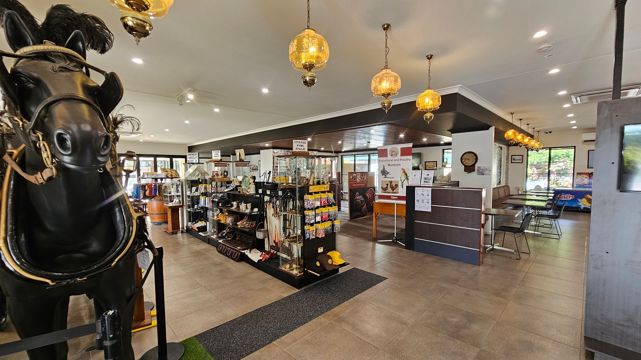 A large black horse statue with a harness is on the left. In the centre, there are shelves displaying various items for sale, including kitchenware and gardening tools. A sign for an "Avicultural and Poultry Museum" is visible. On the right, there are tables and chairs arranged in a dining area. The ceiling features hanging amber-coloured lamps, and the floor is tiled. Large windows in the background allow natural light to enter.