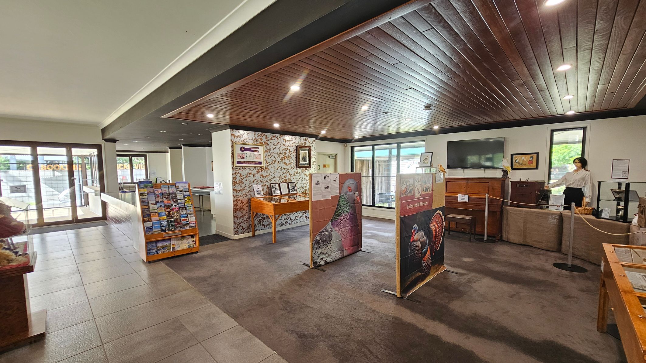 A spacious indoor area with a mix of tiled and carpeted flooring. On the left, there are large glass doors leading outside and a stand with brochures. In the centre, there are display boards featuring bird images. The right side has a mannequin dressed in a white shirt and black skirt, standing behind a rope barrier. The walls have framed pictures and a large TV. The ceiling is wooden with recessed lighting.