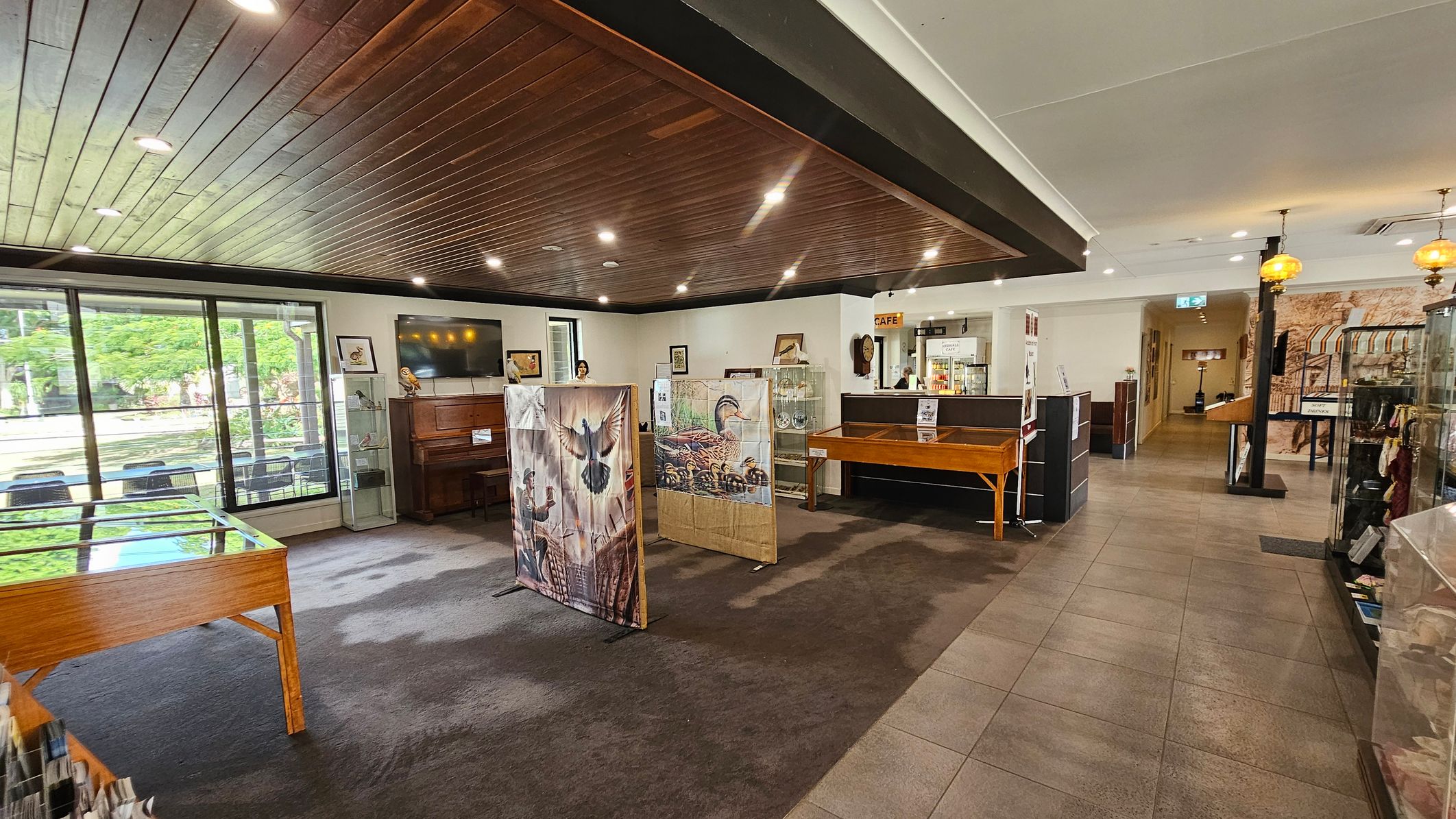 A spacious interior with a wooden ceiling and a mix of carpet and tiled flooring. On the left, large windows offer a view of greenery outside. A wooden piano and display cases with various items are visible. In the centre, there are art panels featuring bird illustrations. To the right, a café counter is present, with a hallway leading further inside. Hanging lights and additional display cases are also on the right.