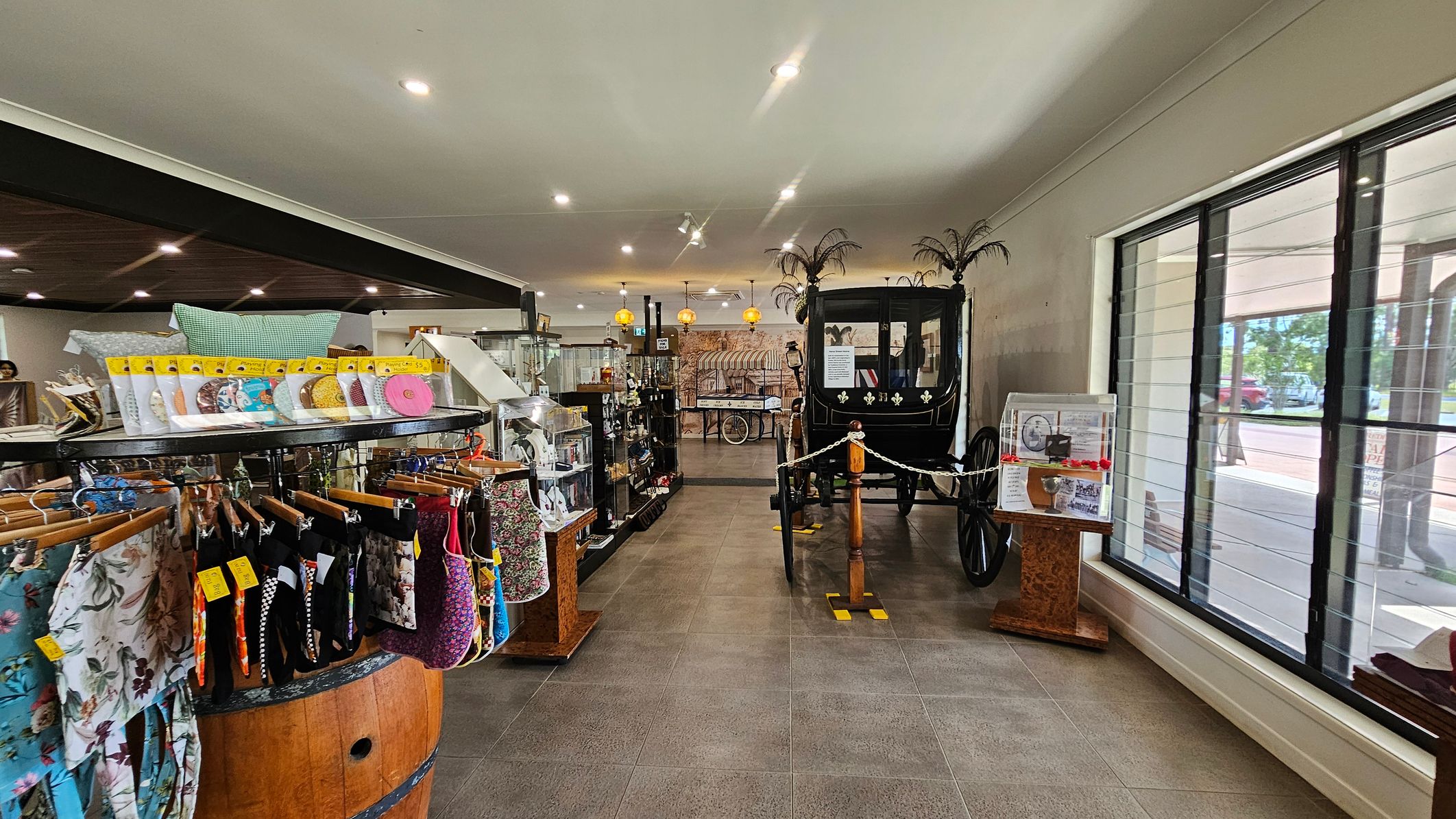 A shop interior with a display of colourful fabric items on hangers to the left, set on a wooden barrel. In the centre, a black vintage carriage with decorative elements is roped off. To the right, large windows allow natural light in, with a stand displaying information. The floor is tiled, and the ceiling has recessed lighting. In the background, shelves with various items and hanging lights are visible.