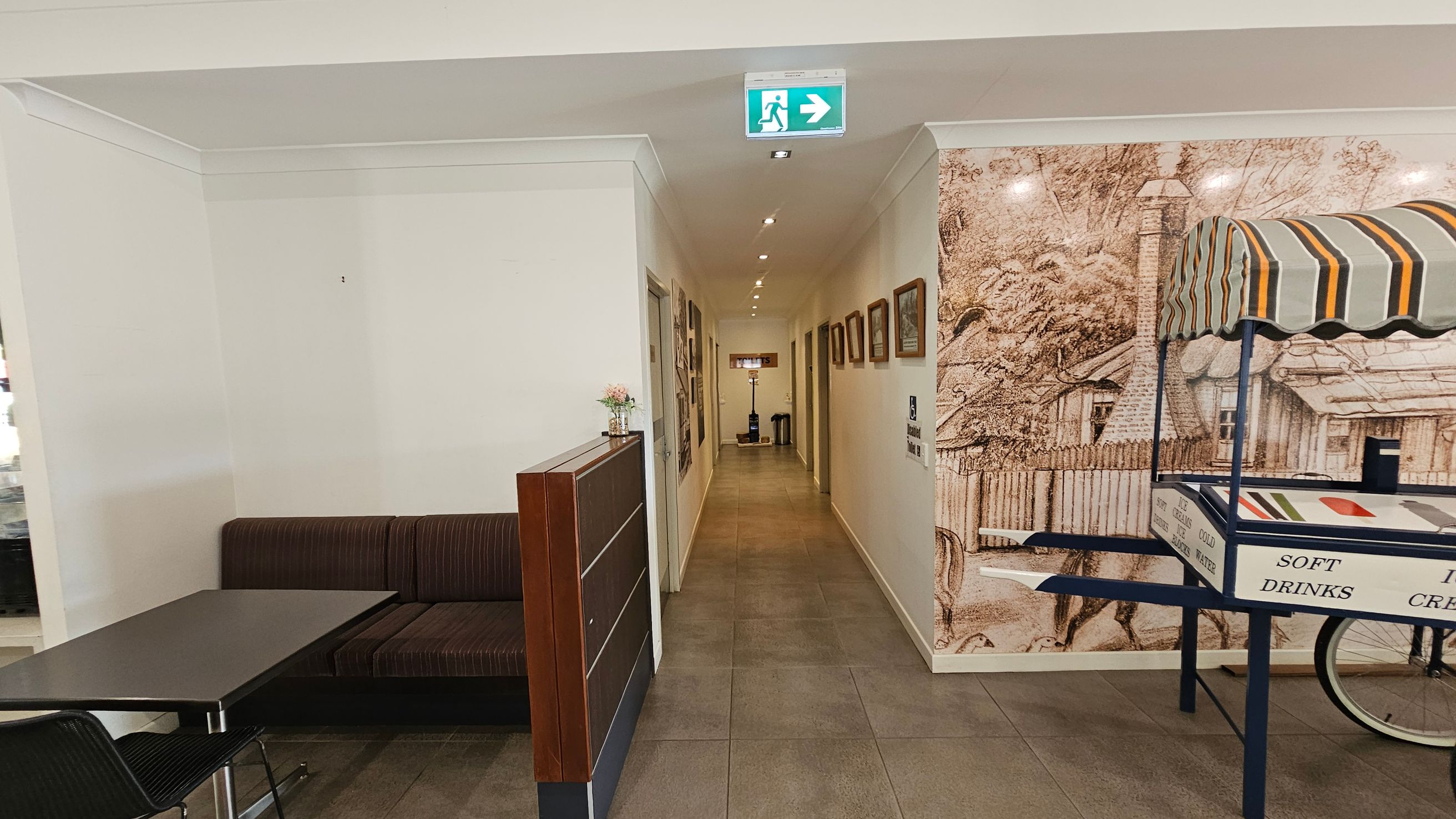 A hallway with a tiled floor and a ceiling with recessed lighting. On the left, there's a seating area with a dark cushioned bench and a black table. A small counter with a flower vase is nearby. On the right, a vintage-style cart with a striped canopy displays signs for soft drinks and ice cream. The wall behind the cart features a large sepia-toned mural of a rustic scene. An exit sign is visible above the hallway.