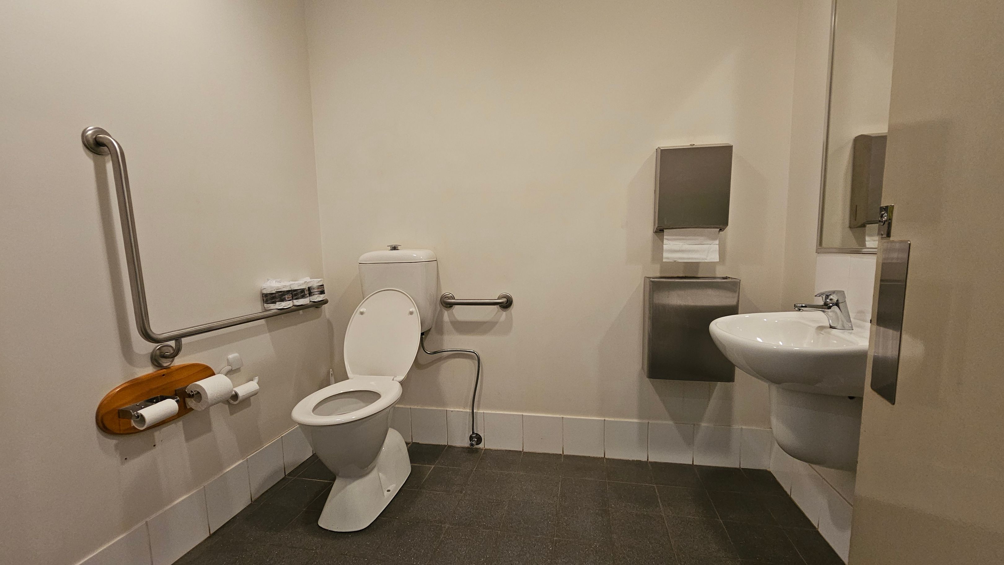 A bathroom with a white toilet on the left, featuring a metal grab bar and a wooden toilet paper holder with two rolls. Above the toilet, there are small bottles on a shelf. On the right, a white sink with a metal tap is mounted on the wall, accompanied by a metal paper towel dispenser. The floor is covered with dark tiles, and the walls are light-coloured. A partially open door is visible on the right side.