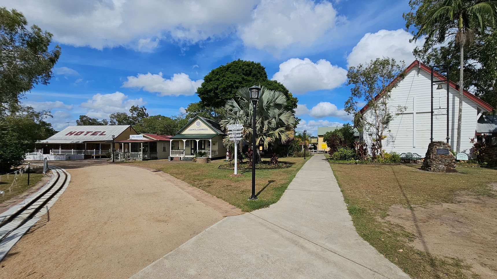 A rural scene with a dirt path on the left leading to a building marked "HOTEL" with a metal roof and a verandah. Next to it is a green building labelled "COUNCIL CHAMBERS" with a gabled roof. A paved path runs through the centre, flanked by a lamp post and a signpost. On the right, a white wooden building with a cross on its facade is visible, surrounded by trees and a small garden. The sky is partly cloudy.