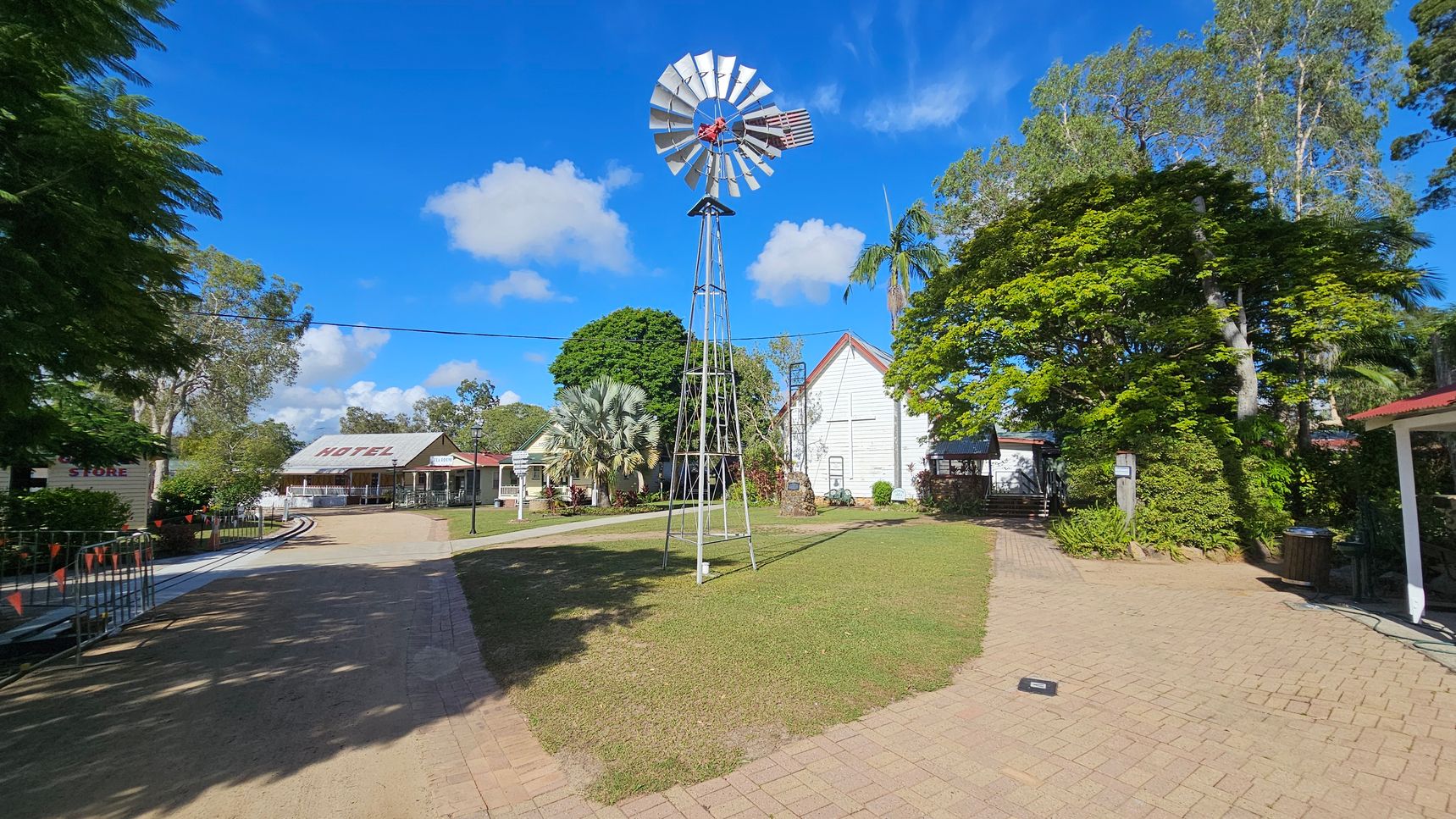 A scene featuring a tall metal windmill in the centre, surrounded by a grassy area. To the left, there's a building with a sign reading "HOTEL" and another structure marked "STORE." The right side has a white building with a red roof, partially obscured by lush green trees. The foreground consists of a paved path, and the background includes more trees and a clear blue sky.