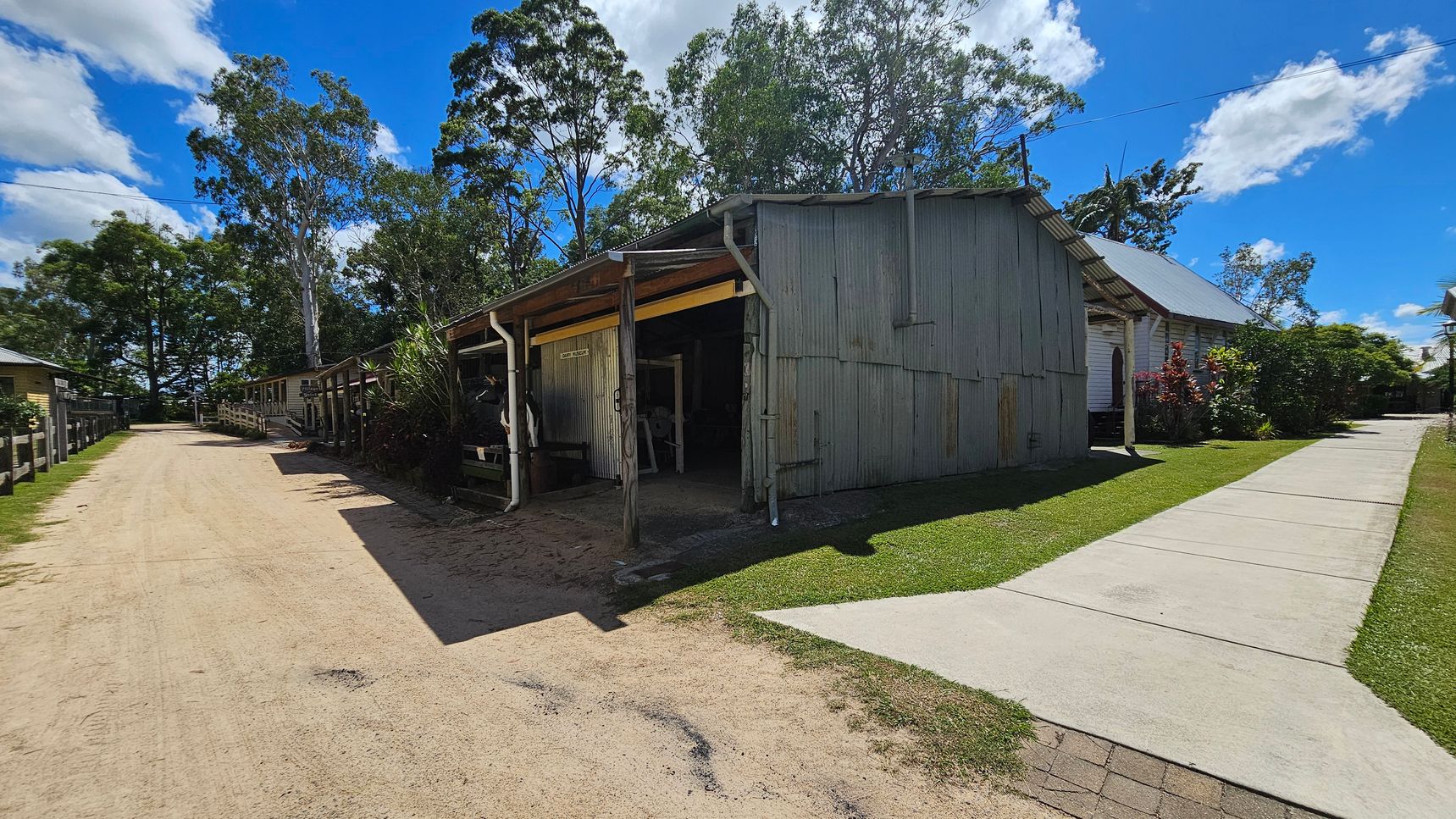 A rustic, weathered wooden building with a corrugated metal roof is on the right, surrounded by green grass and a concrete path. On the left, a dirt road runs alongside a series of similar structures, bordered by a wooden fence. Tall trees with dense foliage are visible in the background, providing shade and a natural backdrop. The scene has a rural, historical feel with a mix of natural and man-made elements.