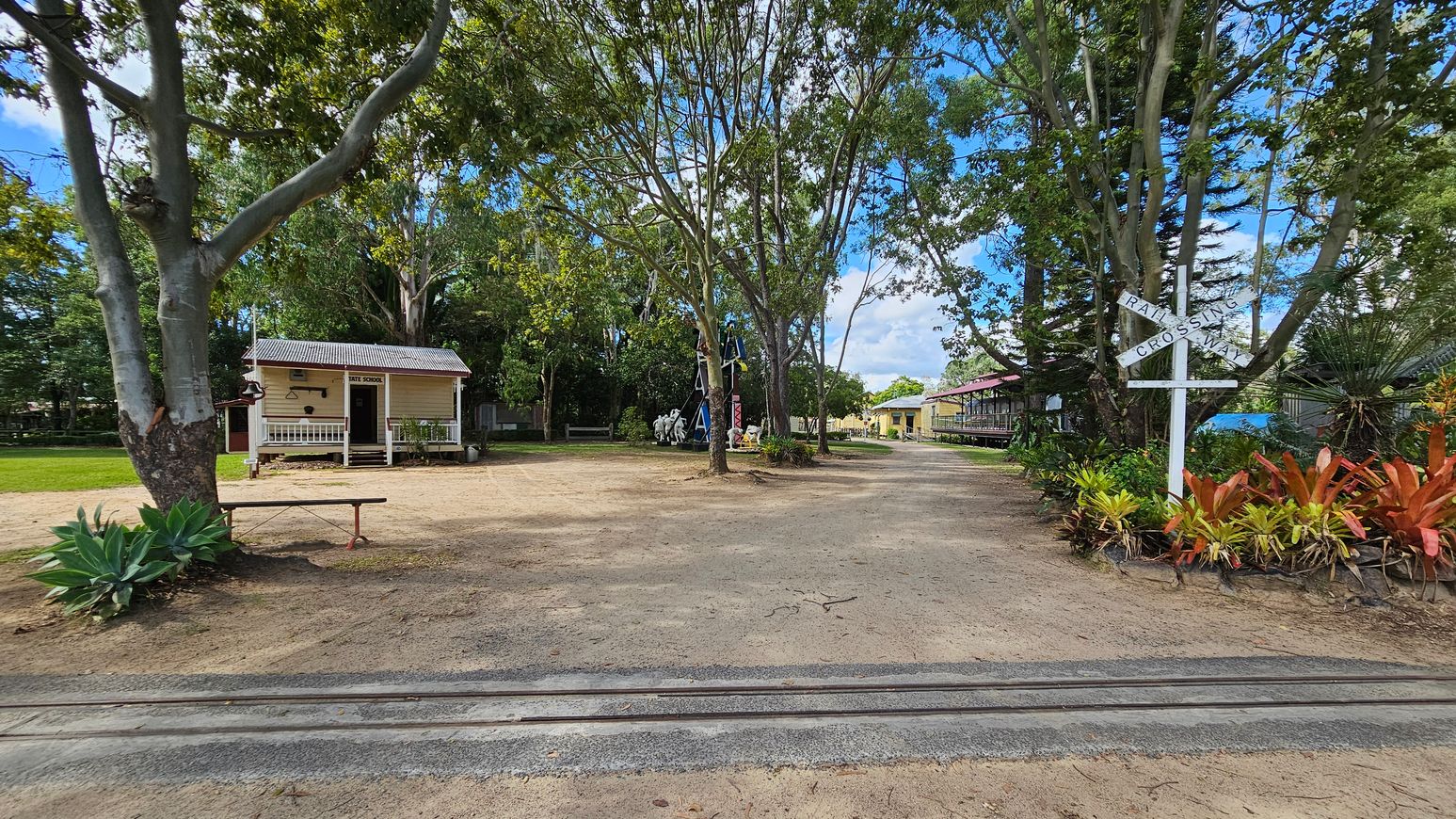 A dirt path runs through the centre, flanked by trees. On the left, a small wooden building with a porch is labelled "State School". On the right, a white railway crossing sign stands amidst colourful plants. In the background, a larger building is visible, partially obscured by trees. The ground is a mix of dirt and sparse grass, and there are railway tracks in the foreground.