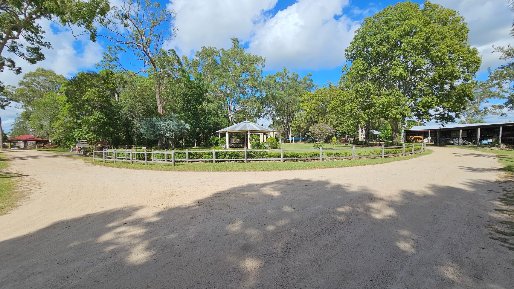 A dirt path curves around a central grassy area with a small gazebo. The gazebo is surrounded by a low wooden fence. On the left, there are trees and a small building with a red roof. On the right, a large tree provides shade, and a structure with open sides houses various equipment. The background is filled with dense greenery and tall trees under a partly cloudy sky.
