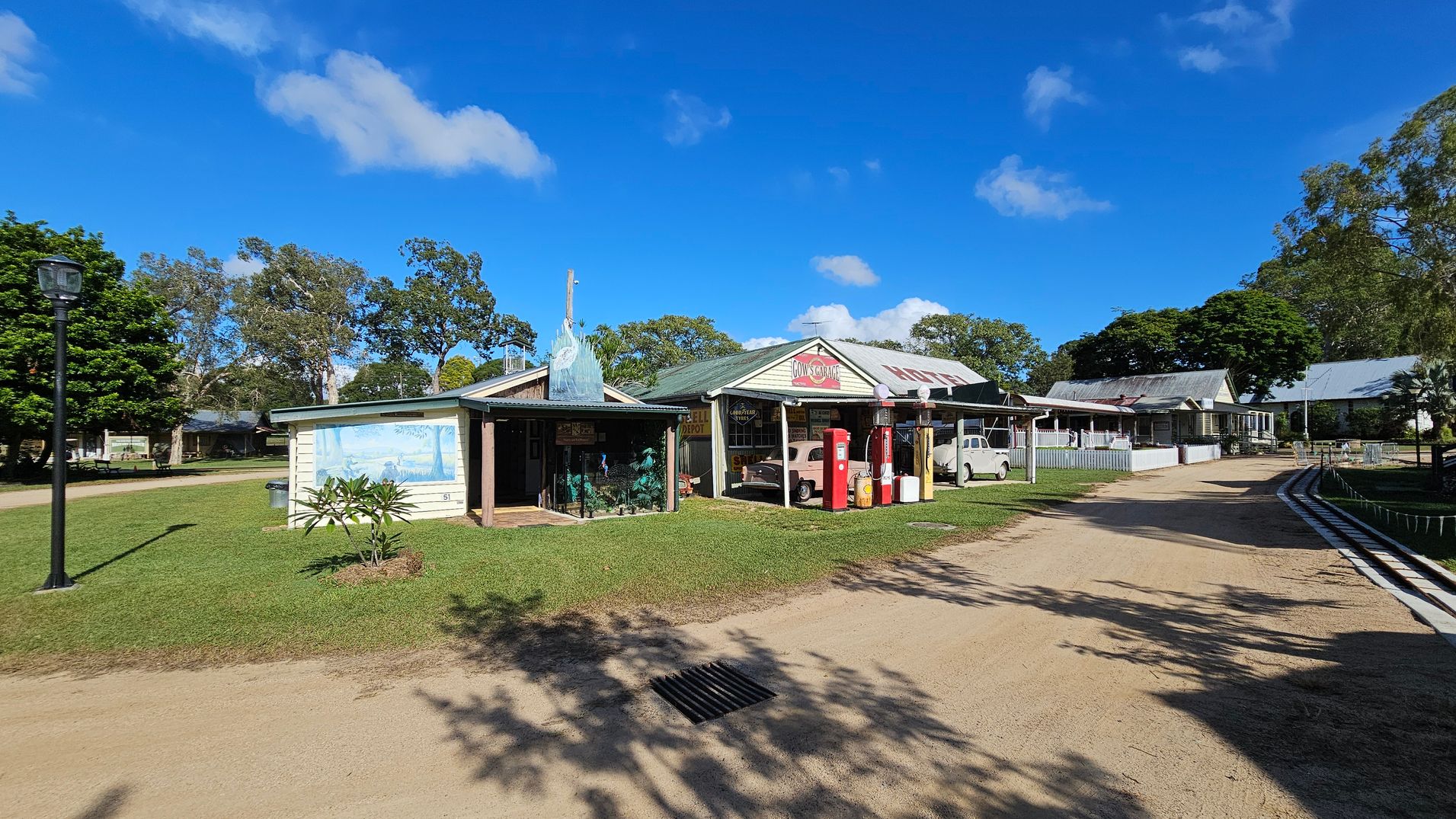 A rural scene with a dirt path running through the centre. On the left, a small building with a mural and a lamp post. In the middle, a larger building with vintage petrol pumps and a classic car under a sign. On the right, more buildings with corrugated roofs and a grassy area. Trees are scattered throughout the background, and the sky is clear with a few clouds.