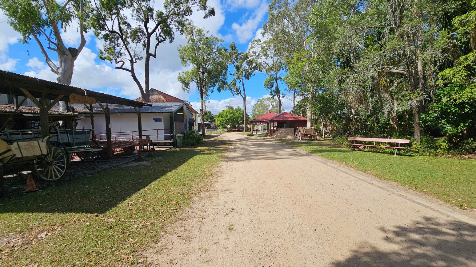 A dirt path runs through the centre, flanked by grass. On the left, there's a wooden structure with a roof, housing old wagons. To the right, a red-roofed shelter and a wooden bench are visible, surrounded by trees and greenery. Tall trees line the path, casting shadows on the ground.