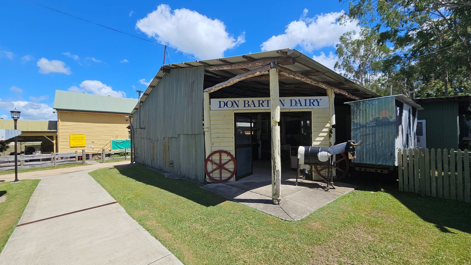 A rustic building with a corrugated metal roof and walls, labelled "Don Barton Dairy," stands on the right. A model cow is positioned outside, near a wooden fence. On the left, a yellow wooden building with a green roof is visible, featuring a sign and a fenced area. A paved path runs through the grass, leading towards the buildings. Trees are in the background, and a lamppost is on the left side of the path.