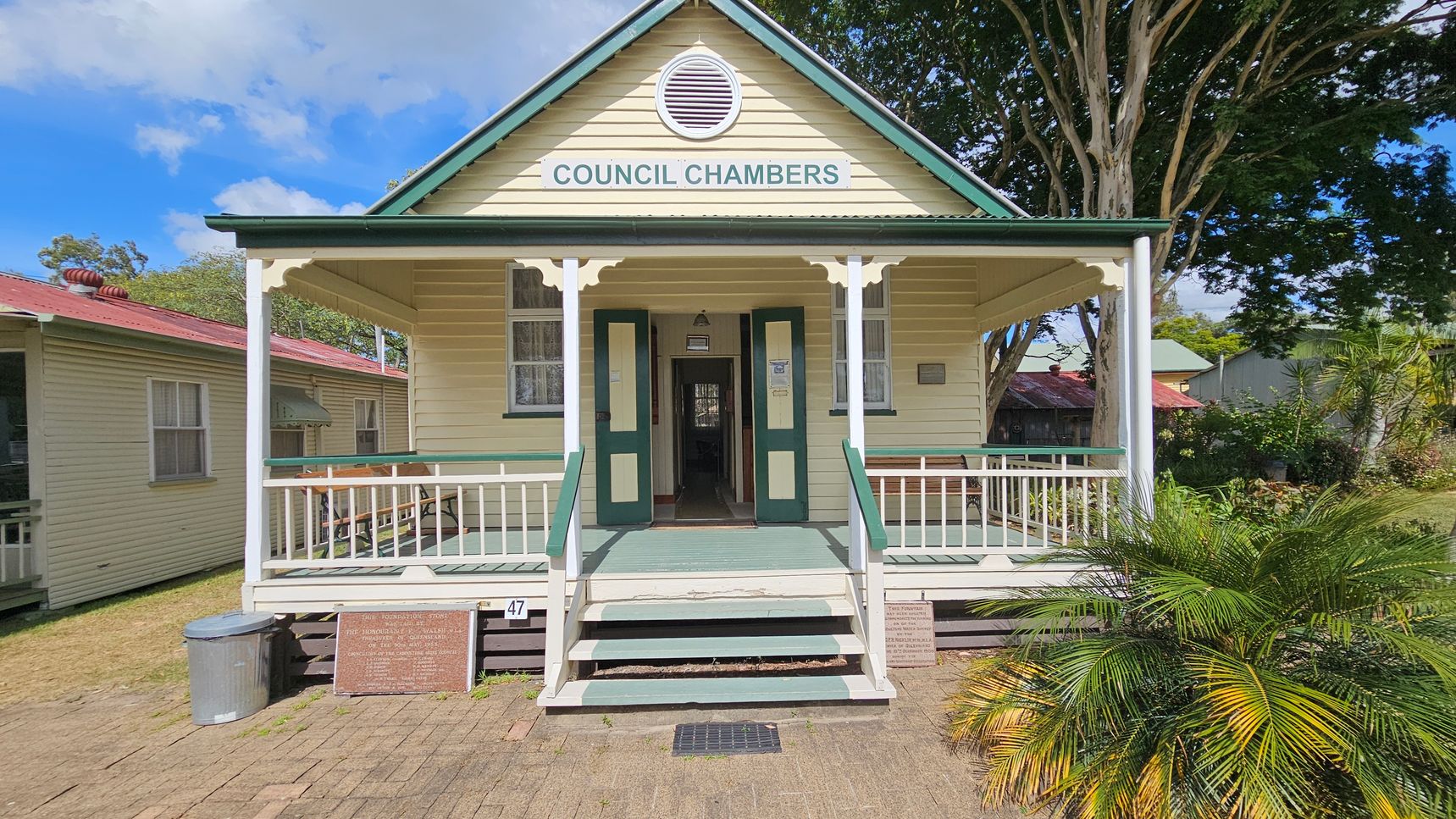 A small, single-storey building with a gabled roof and a sign reading "Council Chambers" above the entrance. The structure is painted in light colours with green trim. A set of steps leads up to a porch with white railings. On the left, another building with a red roof is partially visible. On the right, there are trees and shrubs, including a palm. The ground is paved with bricks, and a metal bin is placed near the steps.