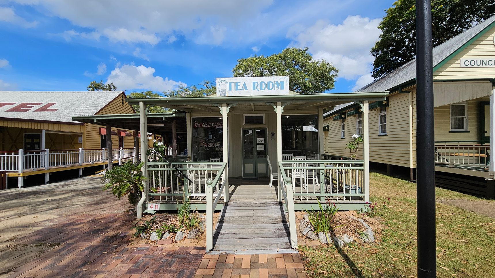 A quaint tea room with a light green exterior and a small porch featuring white chairs. A sign above reads "Tea Room". The building is flanked by two other structures; on the left, a building with a red roof and white railing, and on the right, a cream-coloured building with green trim. The ground is a mix of grass and brick paving. A black lamppost stands to the right, and trees are visible in the background.
