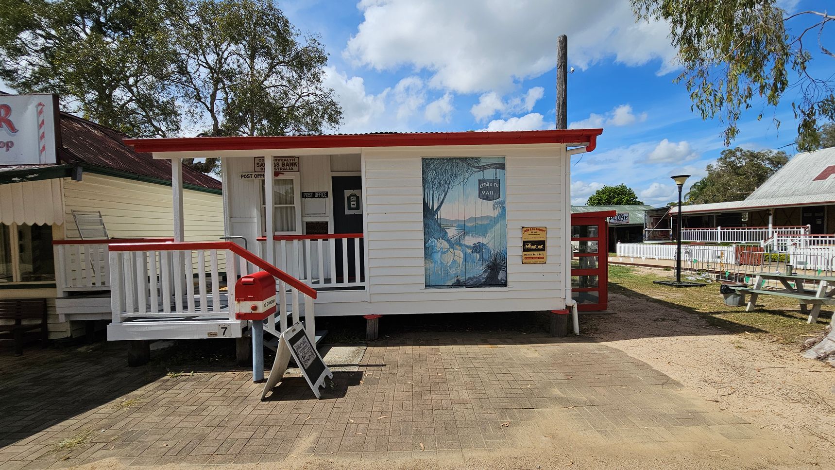 A small white wooden building with a red roof and trim, featuring a porch with white railings and a red handrail. A red post box is mounted near the entrance. A large mural depicting a scenic landscape is on the front wall. To the left, a neighbouring building with a yellow exterior and a red roof is partially visible. On the right, a picnic table and a lamppost are in a grassy area. The ground is a mix of paving and sand, with trees in the background.