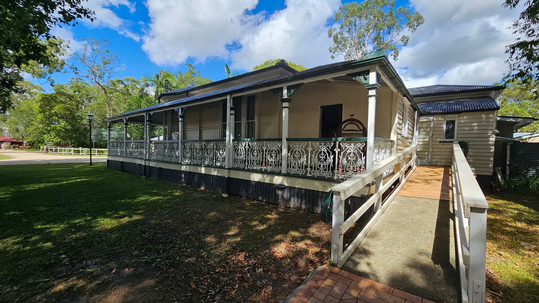 A single-storey house with a decorative metal railing along the veranda. The house has a sloped roof and a wooden ramp leading to the entrance. The exterior is light-coloured with vertical siding. On the left, there's a grassy area with a lamppost and trees in the background. The foreground shows a paved path leading to the ramp. The sky is partly cloudy.