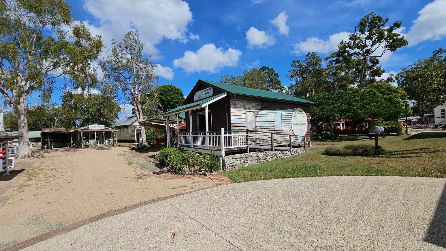 A rustic wooden building with a green roof and a sign reading "Welcome Historical Village" is on the right. It has a small porch with white railings and is surrounded by stonework and greenery. On the left, there is a dirt path leading to another building marked "Tea Room." Tall trees with sparse foliage are scattered throughout the area. The ground is a mix of gravel and grass, and the sky is partly cloudy.
