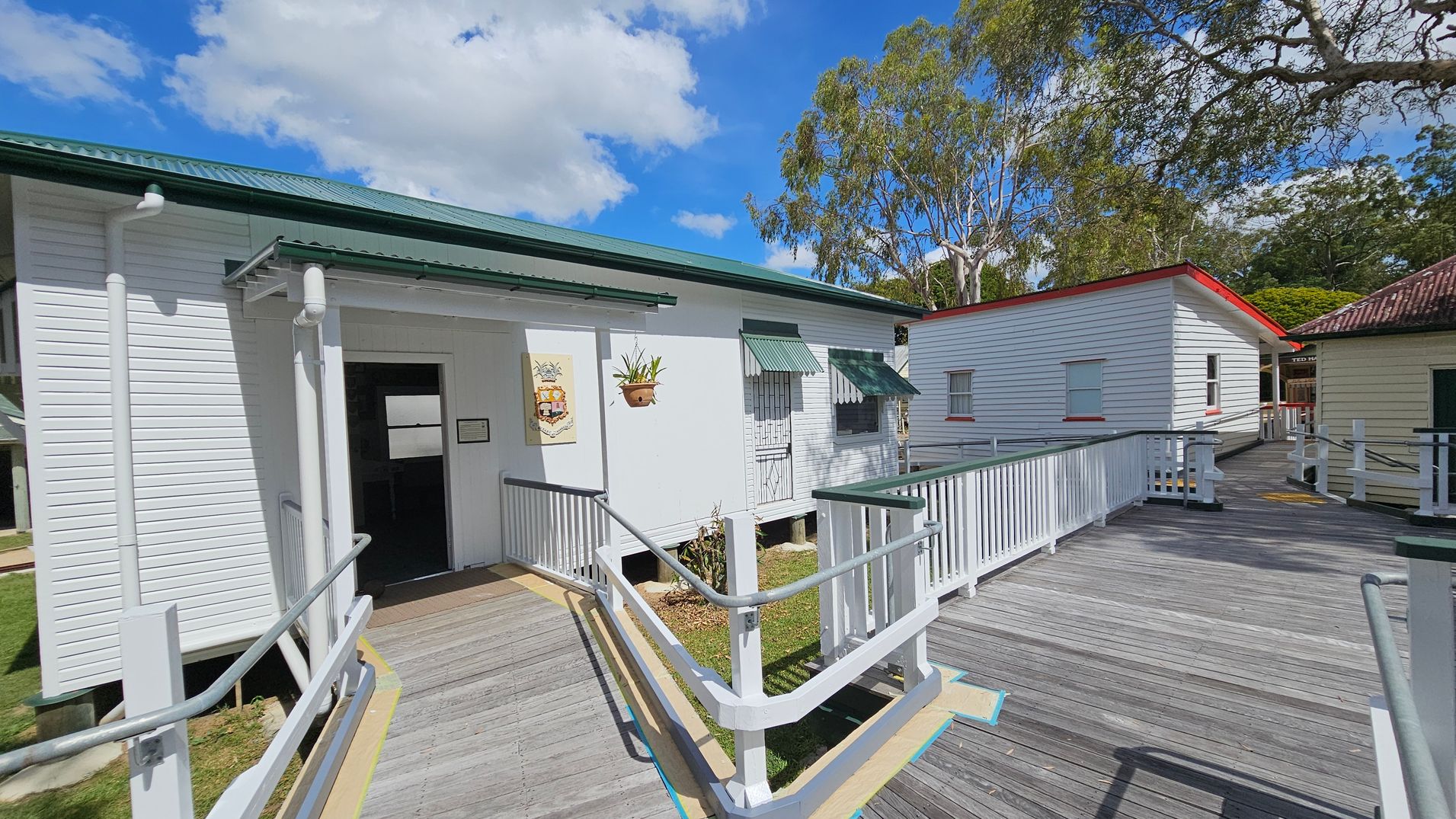 A wooden walkway with white railings leads to a white building with a green roof and awnings on the left. A hanging plant and a decorative sign are near the entrance. To the right, another white building with a red-trimmed roof is visible. Trees with green foliage are in the background, and the sky is partly cloudy. The ground is grassy around the buildings.