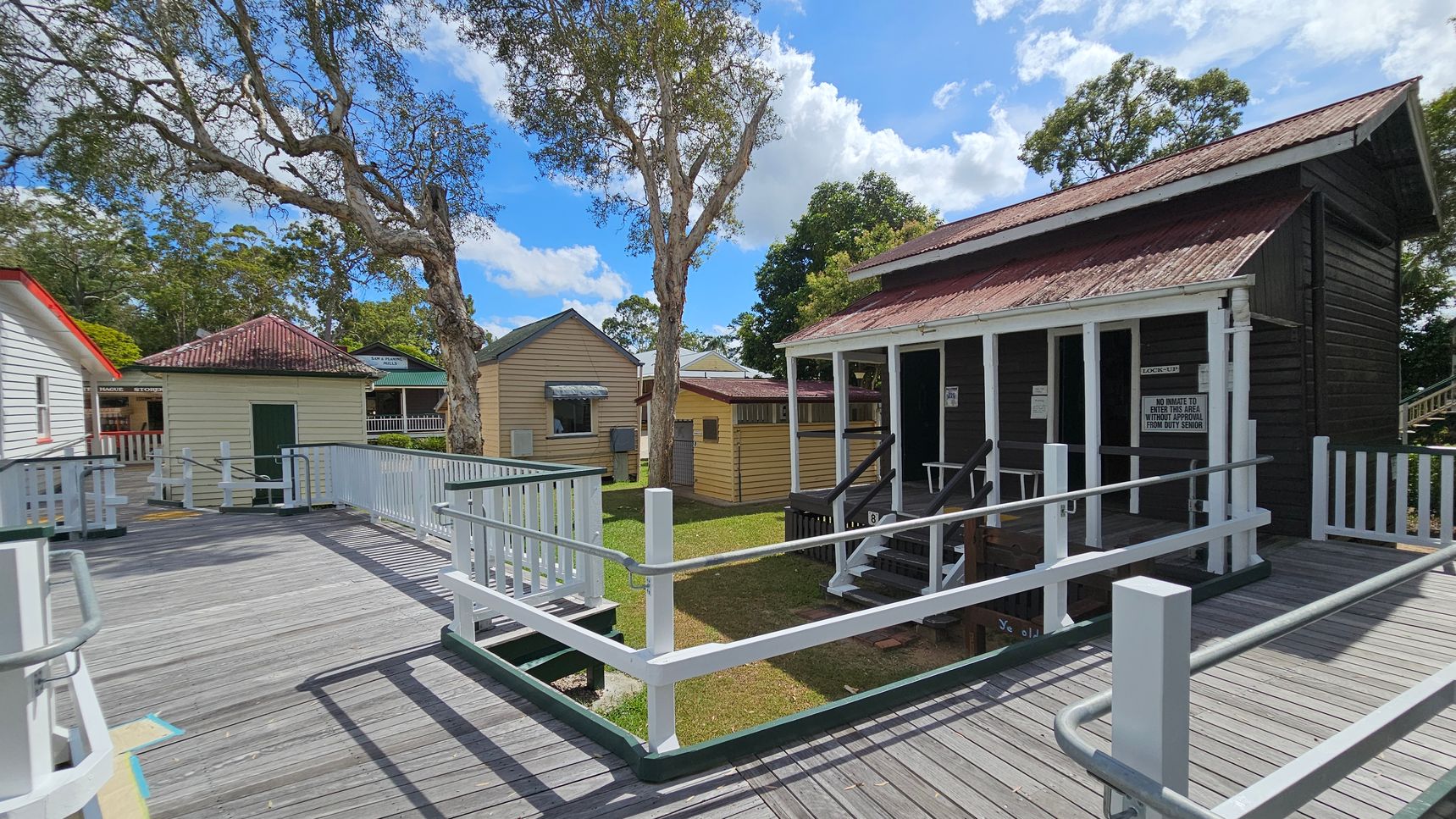 A wooden boardwalk with white railings leads to several small, rustic buildings. On the right, a dark brown building with a corrugated metal roof has a small porch and steps. In the centre, a light yellow building with a similar roof is visible. To the left, a pale green structure with a dark green door stands near a large tree. The background features more trees and a partly cloudy sky. The ground is grassy, and the area appears to be part of a historical or outdoor museum setting.