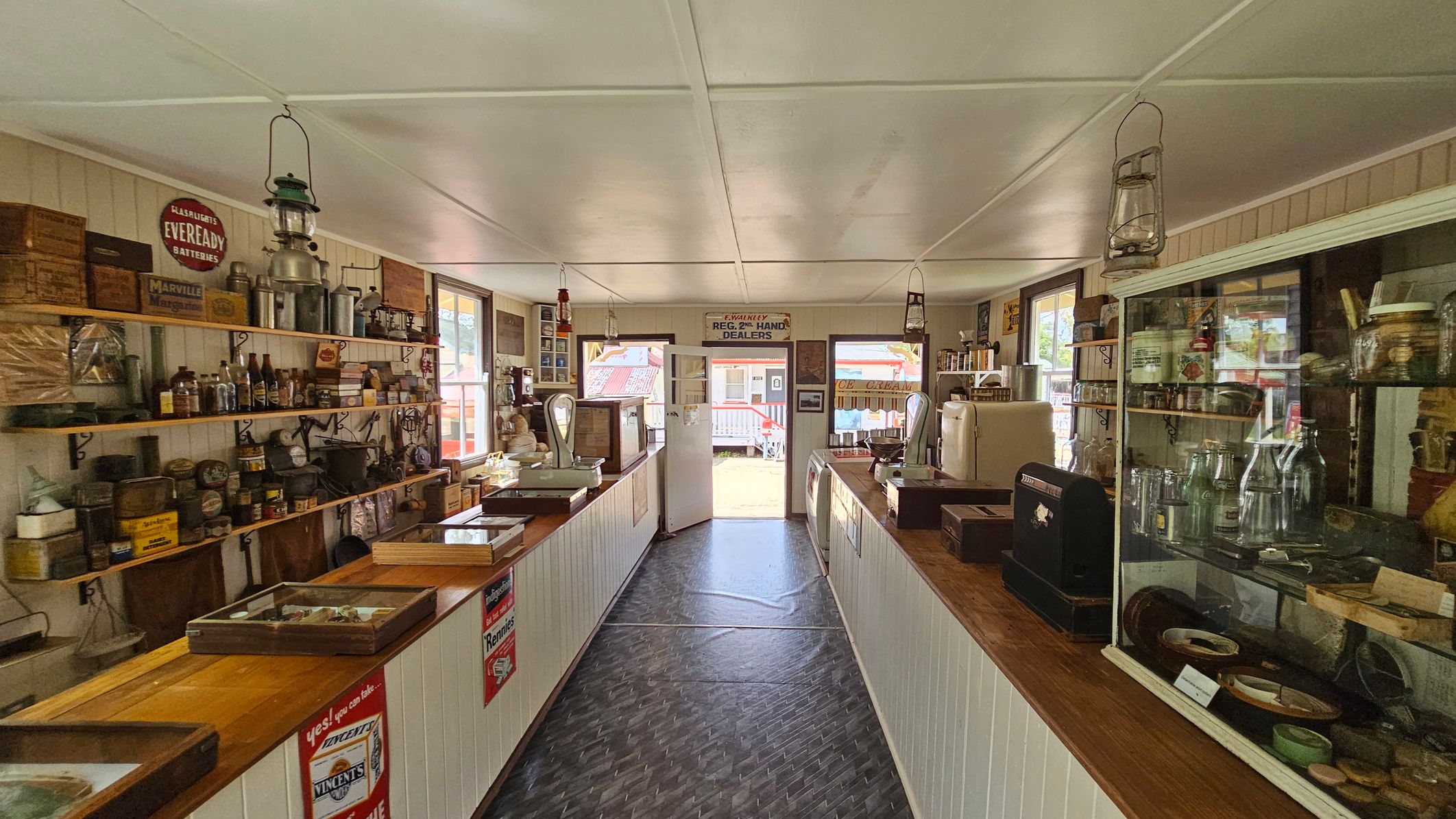 A vintage shop interior with wooden shelves lining both walls, displaying various old-fashioned items like tins, bottles, and lanterns. The left side has a hanging lantern and a sign for Eveready batteries. The right side features a glass display case with jars and a cash register. The floor is dark and patterned, leading to an open door at the back, revealing a glimpse of an outdoor area. The ceiling is white with hanging lanterns.