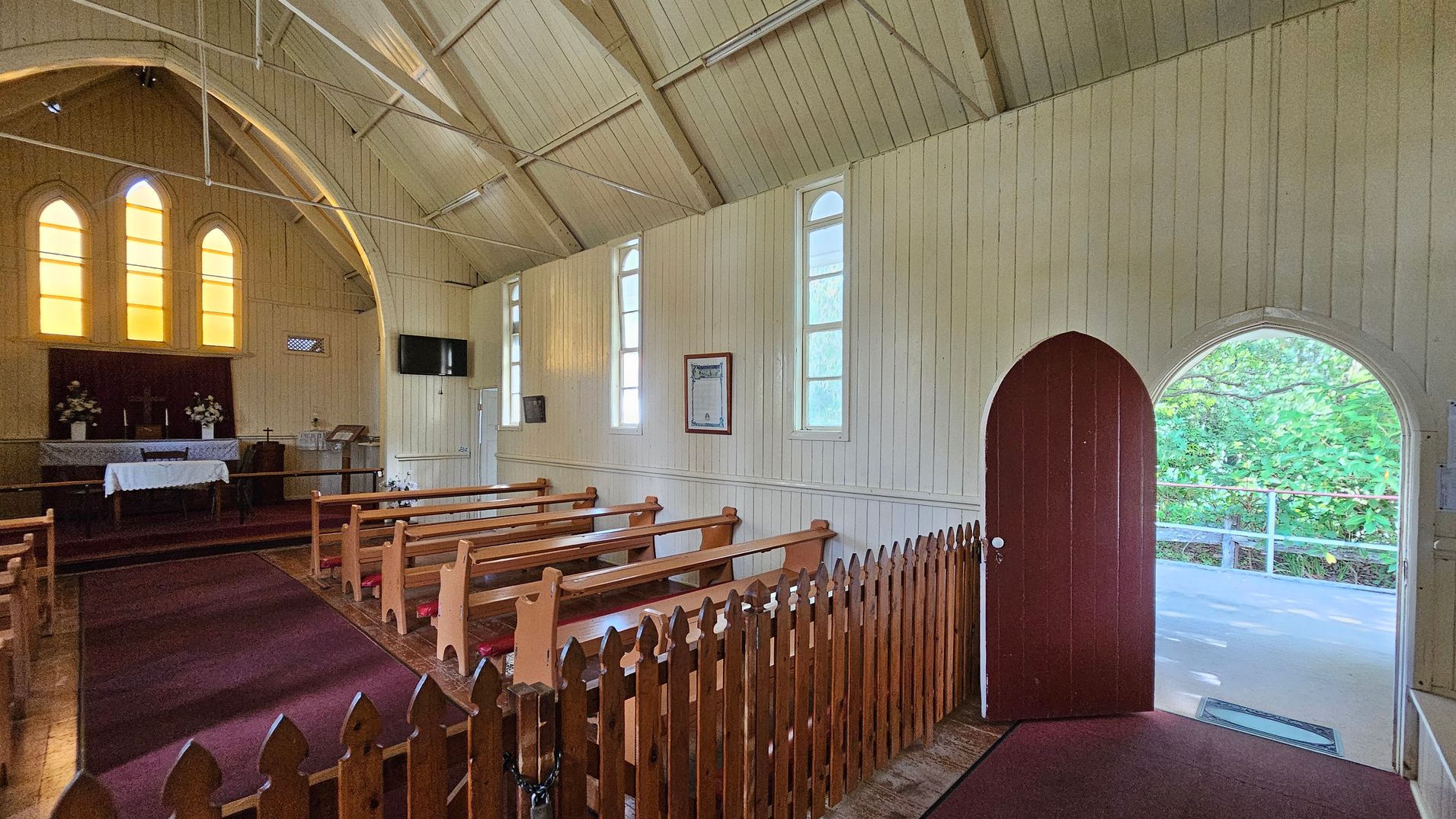 Inside a small church with wooden walls and a vaulted ceiling. On the left, there is an altar with a white cloth and floral arrangements, set against a backdrop of three tall, arched windows with yellow-tinted glass. Wooden pews are arranged in rows, separated by a central aisle with a red carpet. On the right, a wooden arched door is open, leading outside to a view of greenery. A wooden fence lines the aisle, and a flat-screen TV is mounted on the wall.