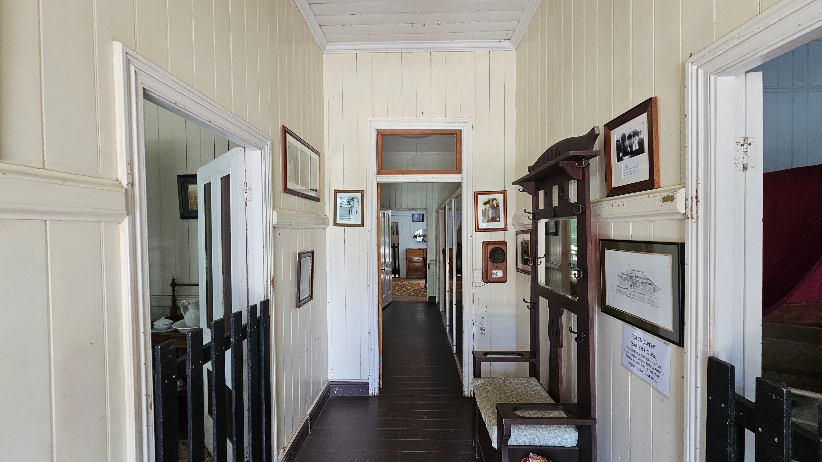A narrow hallway with light-coloured wooden panelled walls and a dark wooden floor. On the left, an open door reveals a room with a bed and a small table. On the right, a wooden coat rack with a mirror and a cushioned bench is visible, along with framed pictures on the wall. At the end of the hallway, another open door leads to a room with a piece of furniture and a window. The ceiling is white and panelled.