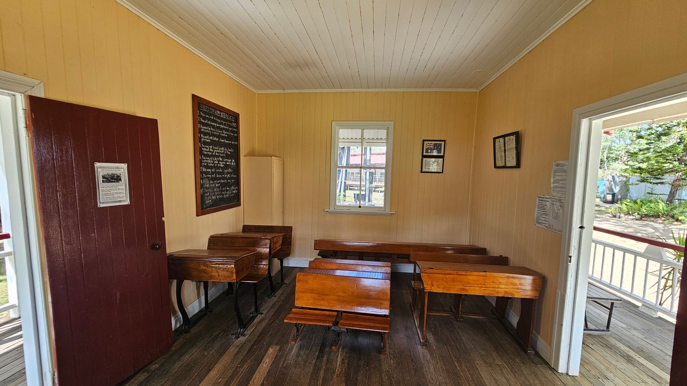 A small, vintage classroom with wooden desks and benches arranged in rows. On the left, a dark red door with a notice leads into the room. A blackboard with writing is mounted on the left wall. In the centre, a window allows natural light to enter, with framed items on the right wall. The floor is wooden, and the ceiling is white with wooden panels. The right side shows an open door leading to a porch with a view of greenery outside.