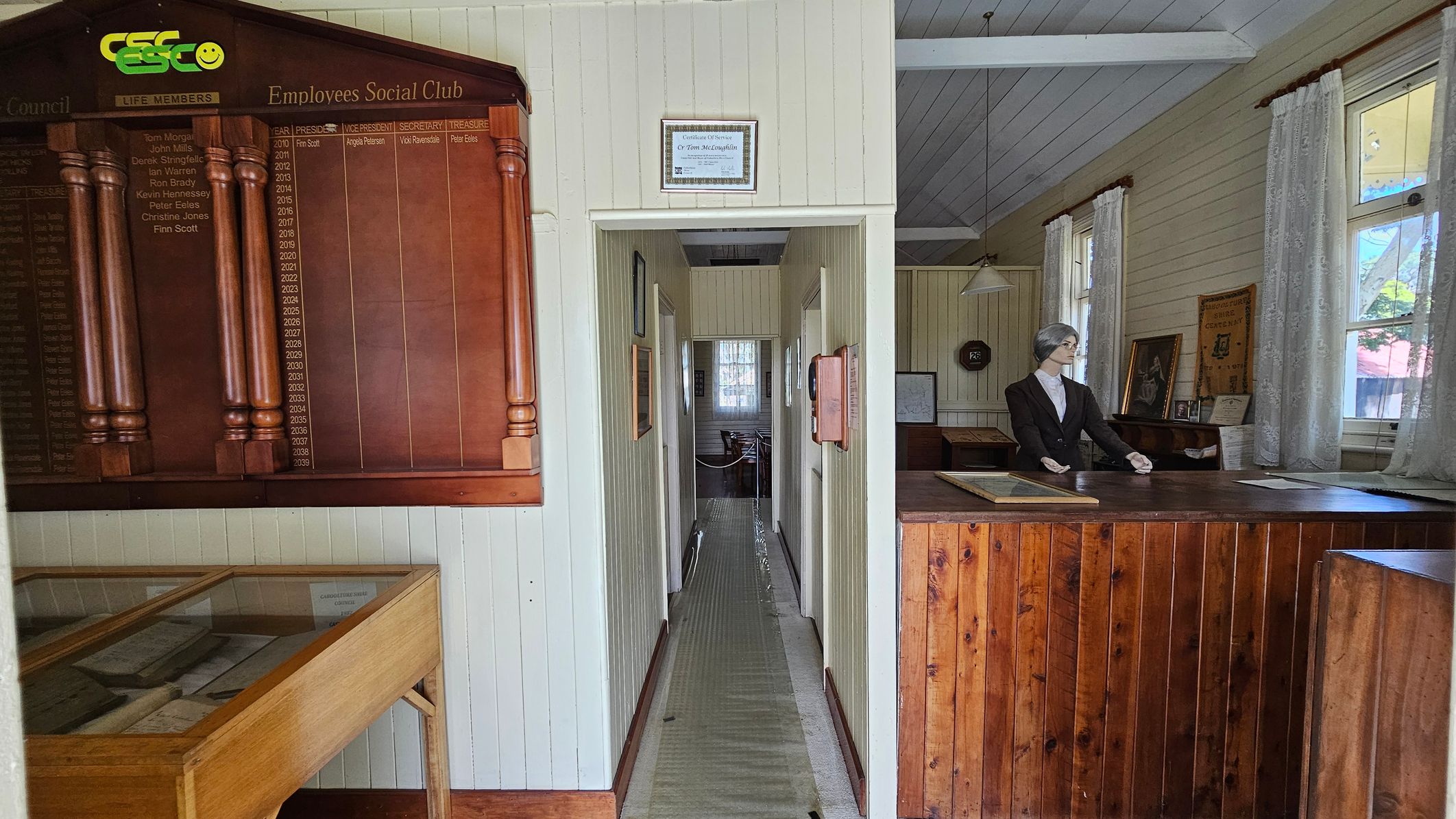 A room with a wooden interior. On the left, a large wooden plaque titled "Employees Social Club" lists names and positions. Below it, a glass display case contains documents. In the centre, a narrow hallway leads to a window. On the right, a mannequin dressed in a suit stands behind a wooden counter. The walls are light-coloured with windows draped in lace curtains. A framed picture and a clock are visible on the right wall. The ceiling is white with exposed beams.