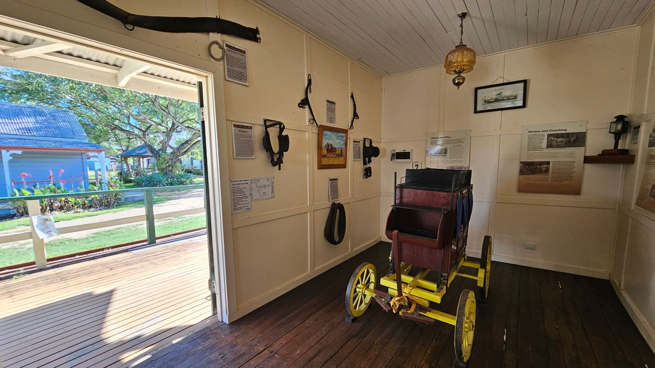 Inside a small room with wooden floors and a white panelled ceiling, a vintage wooden carriage with bright yellow wheels is positioned on the right. The walls are adorned with various horse harnesses and informational plaques. A framed painting and a lantern hang on the walls. An open door on the left leads to a wooden deck outside, where a garden with a large tree and a blue building with a corrugated roof are visible.
