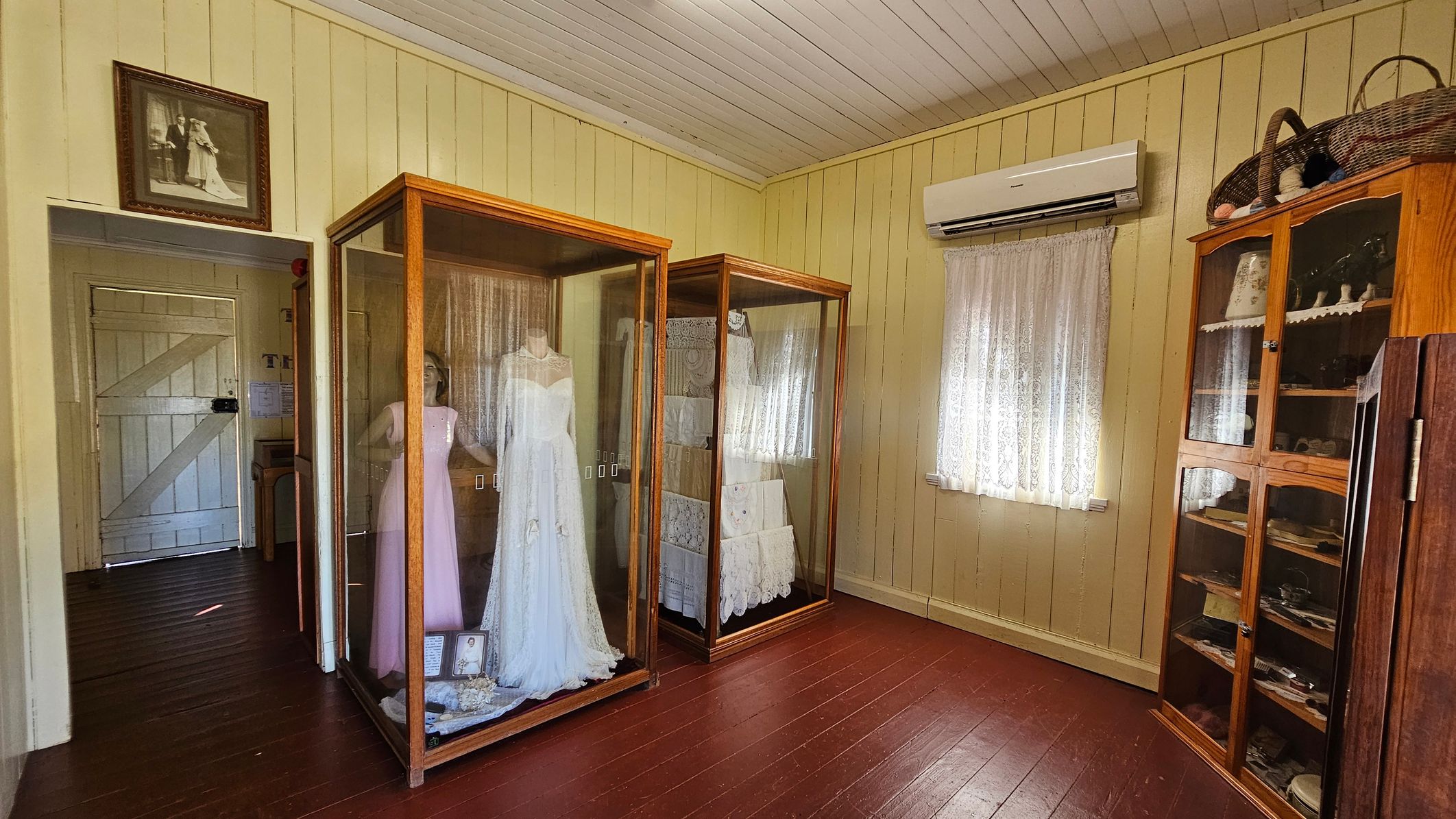 A room with wooden panelled walls and a wooden floor. On the left, there is a doorway with a framed photograph above it. In the centre, two glass display cases contain vintage clothing, including a white dress. On the right, a window with lace curtains is beneath an air conditioning unit. Next to it, a wooden cabinet displays various items, with baskets on top. The ceiling is white and panelled.