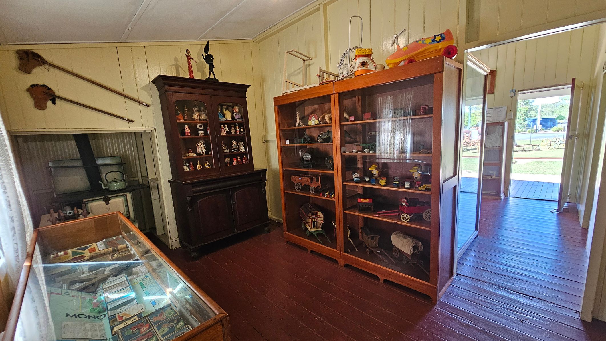 A room with wooden flooring and a white ceiling. On the left, a glass display case contains various documents and items. Above it, two long-handled tools are mounted on the wall. In the centre, a dark wooden cabinet displays small figurines. To the right, a large glass-fronted cabinet showcases vintage toys and models. The room opens to a porch with a view of a grassy area outside.