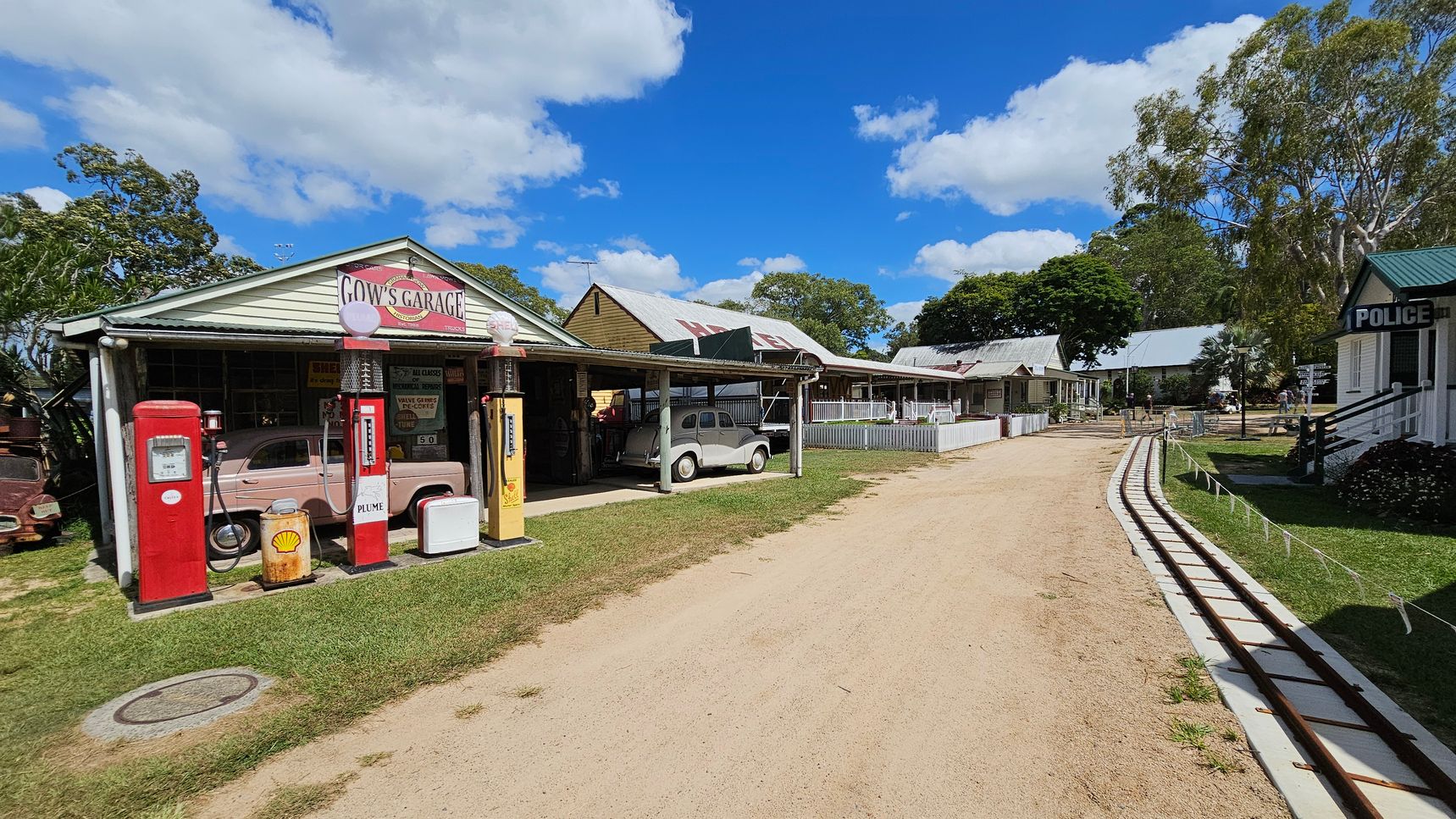A vintage-style scene with a garage on the left, featuring old petrol pumps and a classic car under a canopy. The garage has a sign reading "Gow's Garage." A dirt path runs through the centre, with a small railway track parallel to it on the right. Several old-fashioned buildings line the path, including one marked "Police." Trees and grass are visible throughout, with a clear blue sky above.
