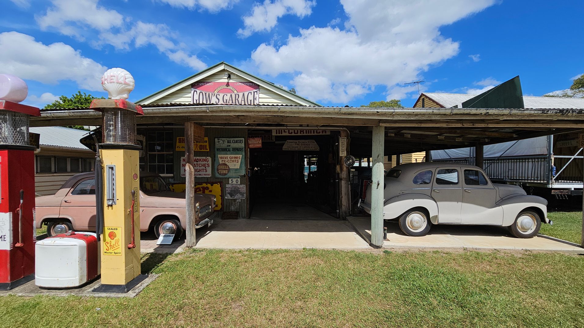 A vintage garage with a sign reading "Gow's Garage" above the entrance. On the left, there are two old petrol pumps, one red and one yellow with a "Shell" logo. A pink classic car is parked beside the pumps. On the right, a grey vintage car is parked under a wooden canopy. The garage has various retro signs and advertisements on the walls. The ground is grassy, and the sky is partly cloudy.