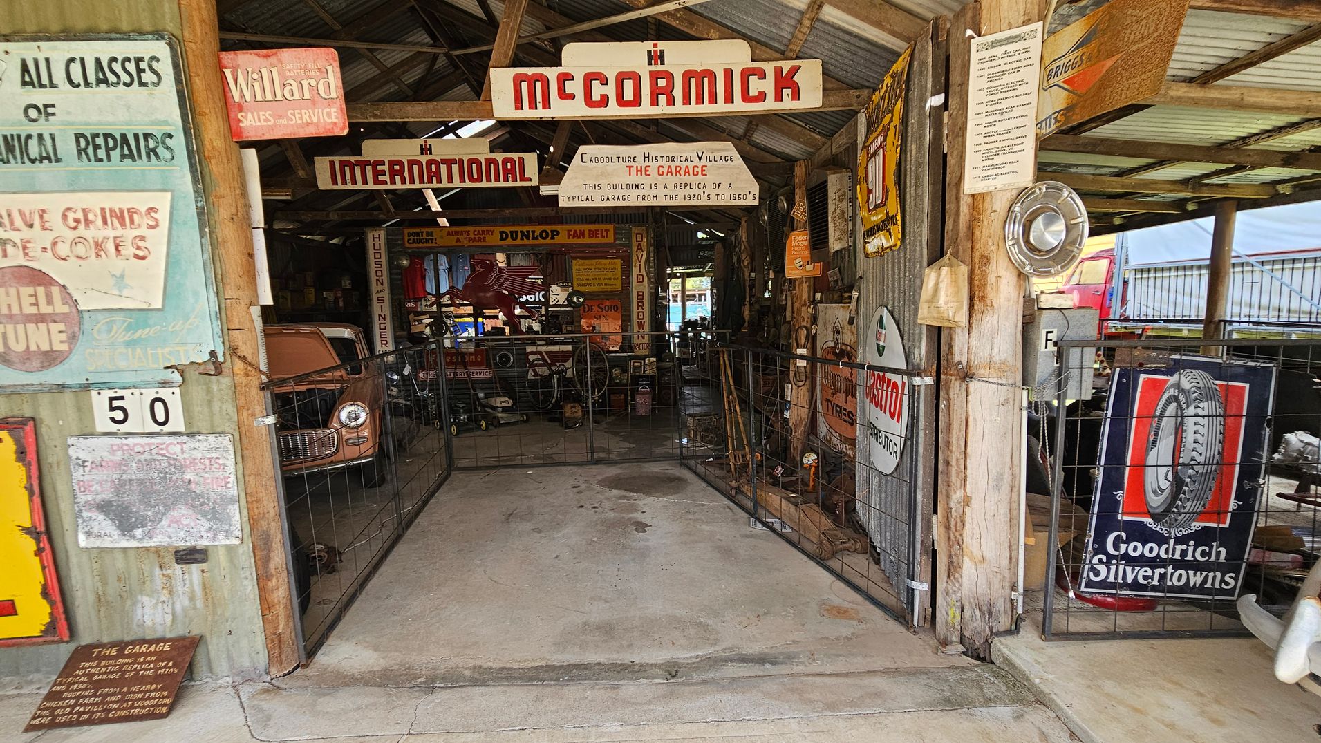 A vintage garage scene with a corrugated metal roof and wooden beams. On the left, various retro signs are displayed, including "Willard Sales and Service" and "All Classes of Mechanical Repairs." In the centre, a sign reads "McCormick" and "International," with a vintage car partially visible. On the right, more signs, including "Goodrich Silvertowns," are attached to the walls. The floor is concrete, and the area is enclosed with metal fencing.
