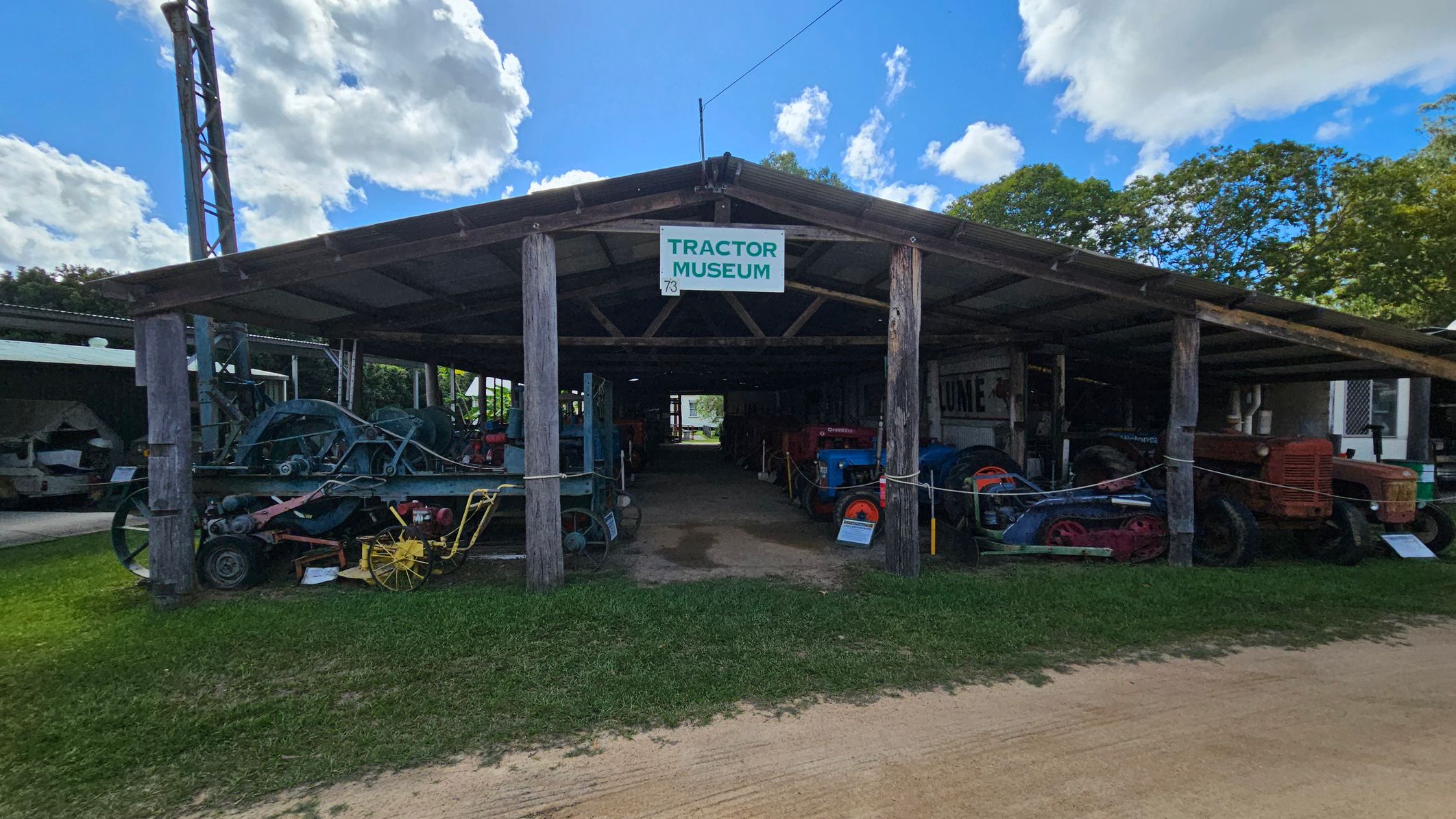 A rustic wooden structure with a sign reading "Tractor Museum" at the entrance. Inside, various vintage tractors are displayed in rows. The building has a simple, open design with a sloped roof supported by wooden beams. The ground is a mix of grass and dirt, and there are trees visible in the background. A tall metal structure is on the left side.