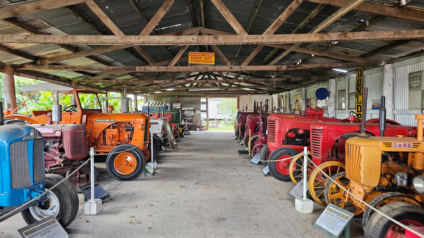 Inside a rustic shed with a corrugated metal roof supported by wooden beams, a collection of vintage tractors is displayed. On the left, there are blue and orange tractors, while on the right, red and yellow tractors are lined up. Each tractor is accompanied by an informational plaque. A sign hanging from the ceiling reads, "Children Please Keep Off Tractors." The floor is concrete, and the walls are lined with various signs and memorabilia. In the background, greenery is visible through the open end of the shed.