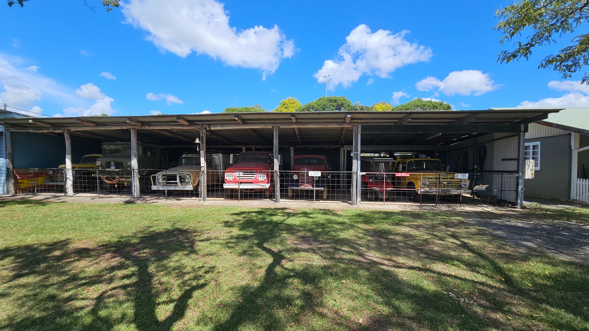A row of vintage vehicles is parked under a large, open-sided shelter with a corrugated metal roof. The vehicles are lined up side by side, each in its own bay, separated by metal posts. The ground is covered with grass, and there are shadows of trees cast on it. In the background, there are trees with green foliage. The shelter has a wire fence in front, and a small building with a window is visible on the far right.