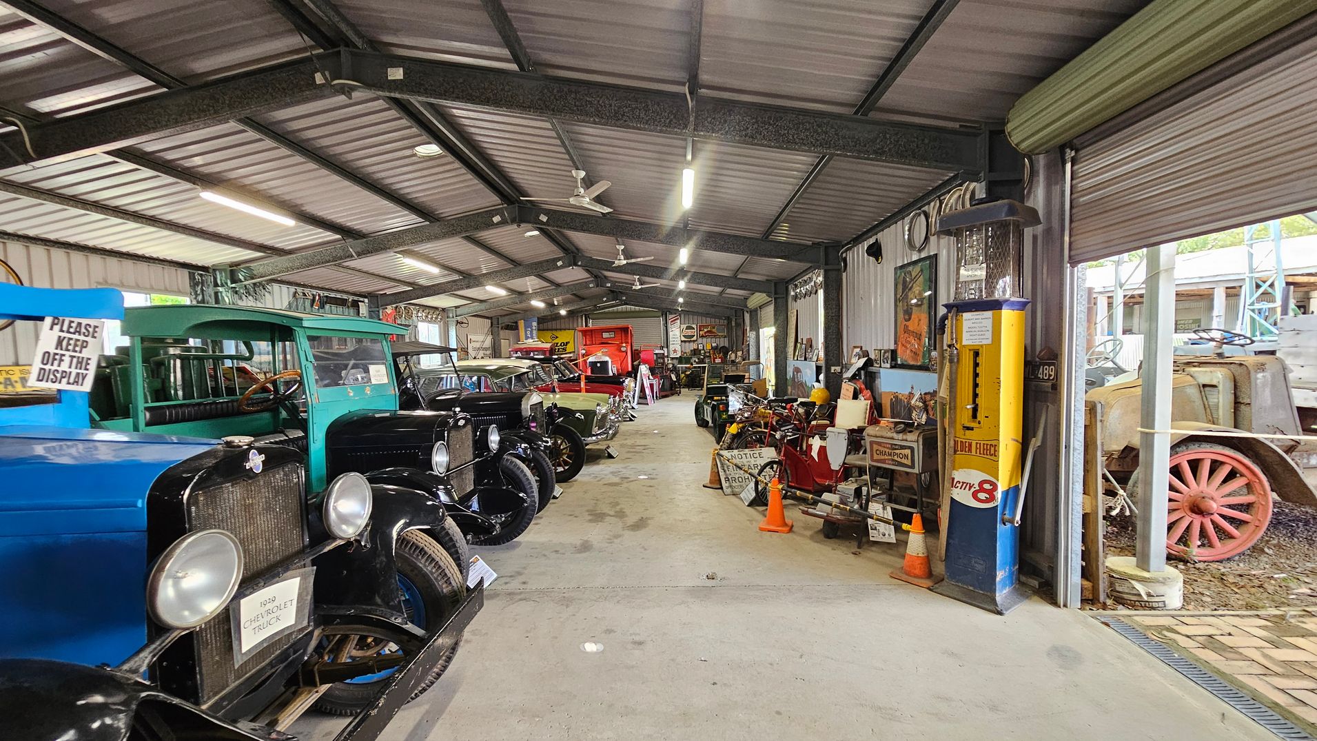 Inside a large, metal-roofed garage, vintage cars are lined up on the left, including a blue and green Chevrolet truck. On the right, various antique items and signs are displayed, along with a yellow and blue petrol pump. The floor is concrete, and the ceiling has exposed beams with fluorescent lights and ceiling fans. A partially open roller door on the right reveals an old vehicle with red wheels outside.