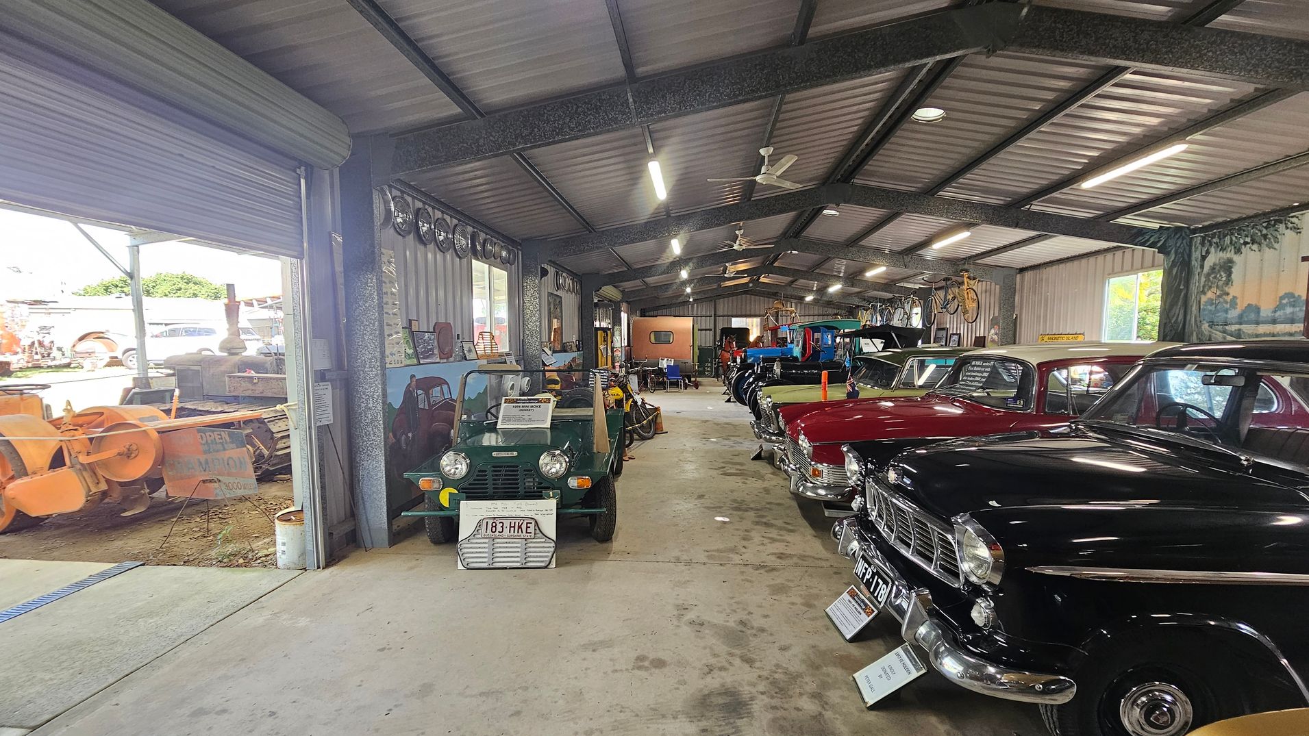 Inside a large metal-roofed garage, classic cars are lined up on the right, including a black car in the foreground and a red one behind it. On the left, a green vintage vehicle is displayed near the entrance. The floor is concrete, and bicycles hang on the wall. The ceiling has exposed beams with fluorescent lights. Outside, visible through the open entrance, are orange farm machinery and greenery.