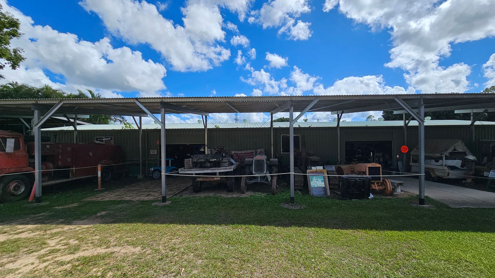 A covered outdoor area with a corrugated metal roof supported by metal beams. Underneath, various machinery and equipment are displayed, including a tractor and other vintage vehicles. A rope barrier separates the display from the grassy foreground. The background features a metal building with open sections revealing more equipment inside. A "STOP" sign is visible on the right. The ground is a mix of grass and dirt.