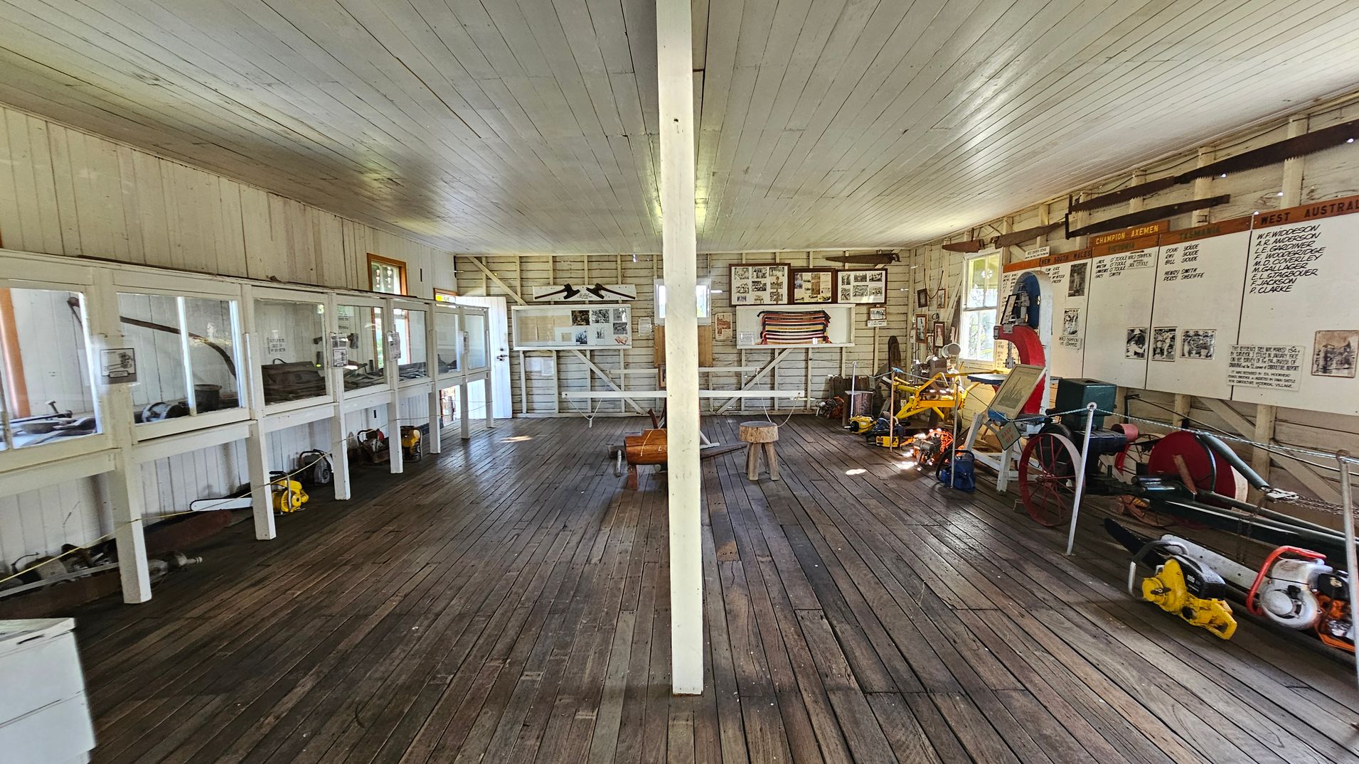 A room with wooden floors and a white wooden ceiling. On the left, there are glass display cases containing various tools and equipment. The right side features a wall with large boards listing names and photographs, along with more tools and machinery on the floor. The back wall has framed pictures and a flag. A central white pillar divides the room, and there are wooden stools and a barrel in the middle. Natural light enters through windows on the right.