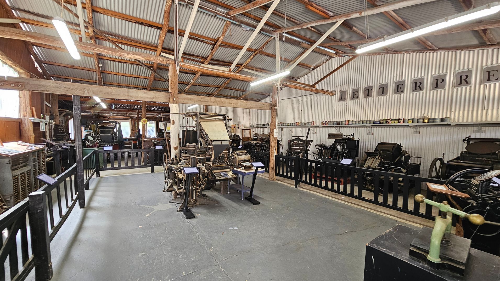 A room with a corrugated metal ceiling and wooden beams. On the left, there are various vintage printing machines behind a black wooden fence. In the centre, a large, intricate printing press is displayed with informational plaques. On the right, more printing equipment is lined up against the wall, which has the word "LETTERPRESS" displayed in large letters. The floor is concrete, and fluorescent lights hang from the ceiling.