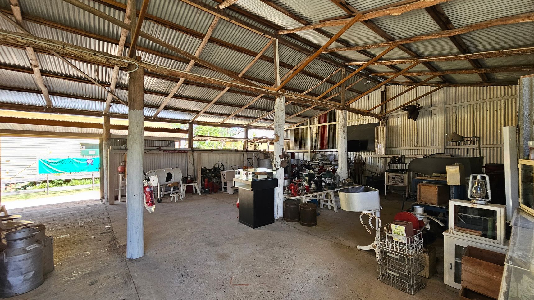 A rustic shed with a corrugated metal roof and wooden beams. On the right, various vintage machinery and equipment are displayed, including a large green machine and a lantern on a white cabinet. In the centre, a black pedestal and a metal basin are visible. On the left, more machinery and a blue banner can be seen through the open side of the shed. The floor is concrete, and the walls are made of corrugated metal.