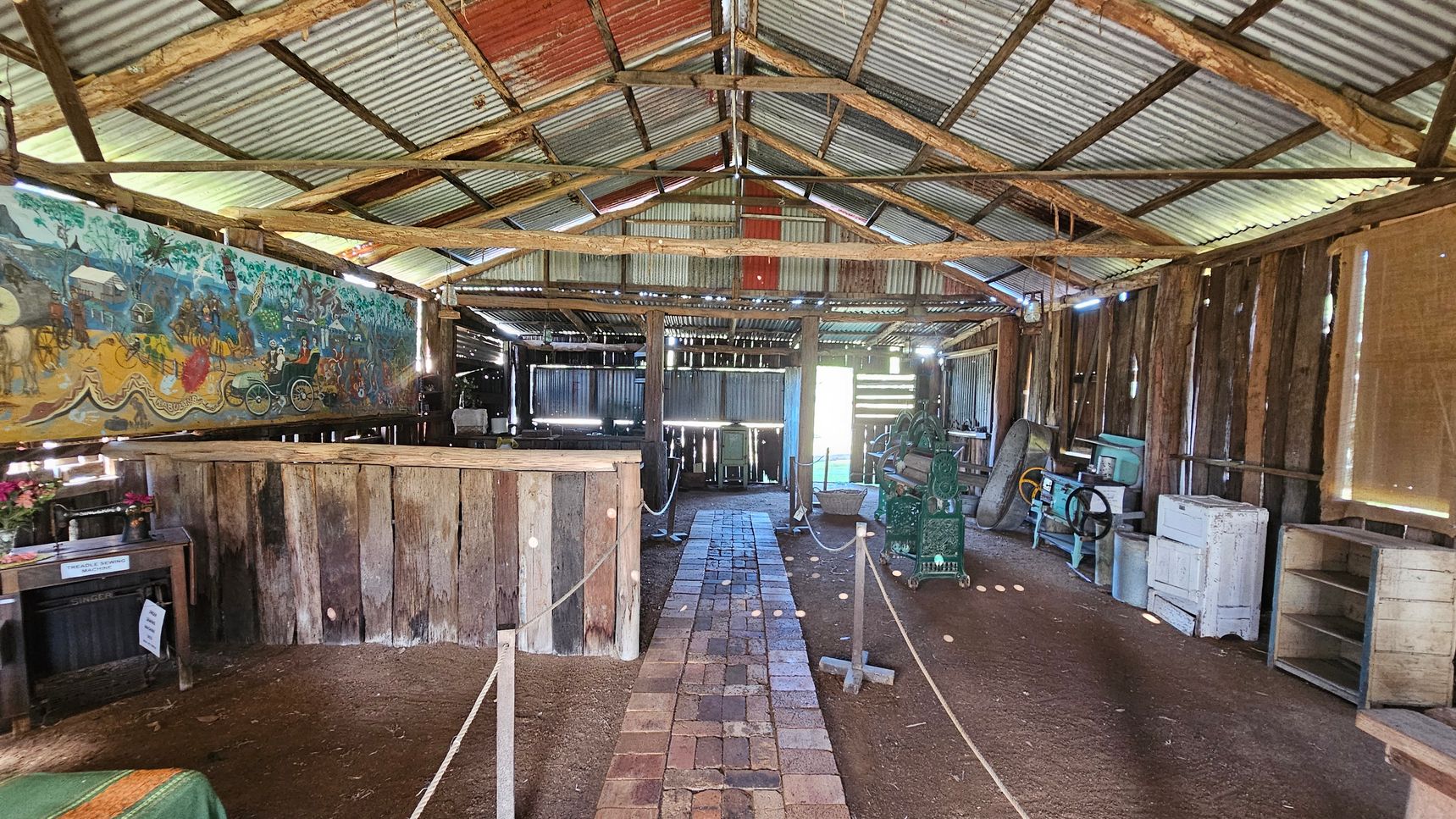 Inside a rustic shed with a corrugated metal roof and wooden beams. The floor is dirt with a central path made of bricks. On the left, a colourful mural decorates the wall above a wooden counter. Various vintage machinery and equipment are scattered throughout, including a green machine on the right. The walls are made of wooden planks, and there are ropes sectioning off areas. The shed is lit by natural light filtering through gaps in the walls and roof.
