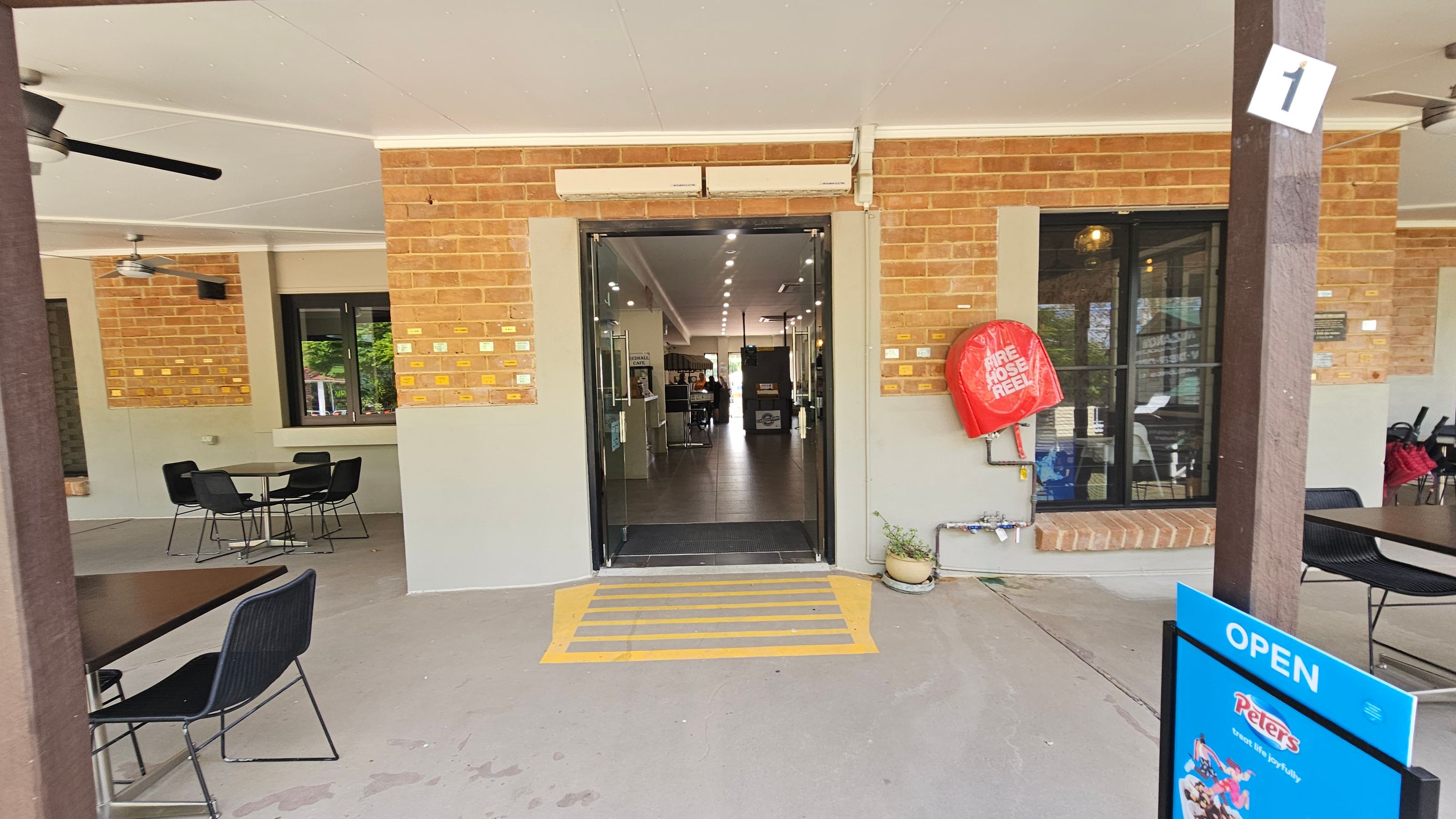 A brick building entrance with a glass door in the centre, leading to an interior with tiled flooring and ceiling lights. On the right, a red fire hose reel is mounted on the wall, and a blue "OPEN" sign is visible. On the left, there are outdoor tables and chairs under a ceiling with fans. The floor is concrete, and there are small plaques on the walls. A wooden post with a number "1" is in the foreground.