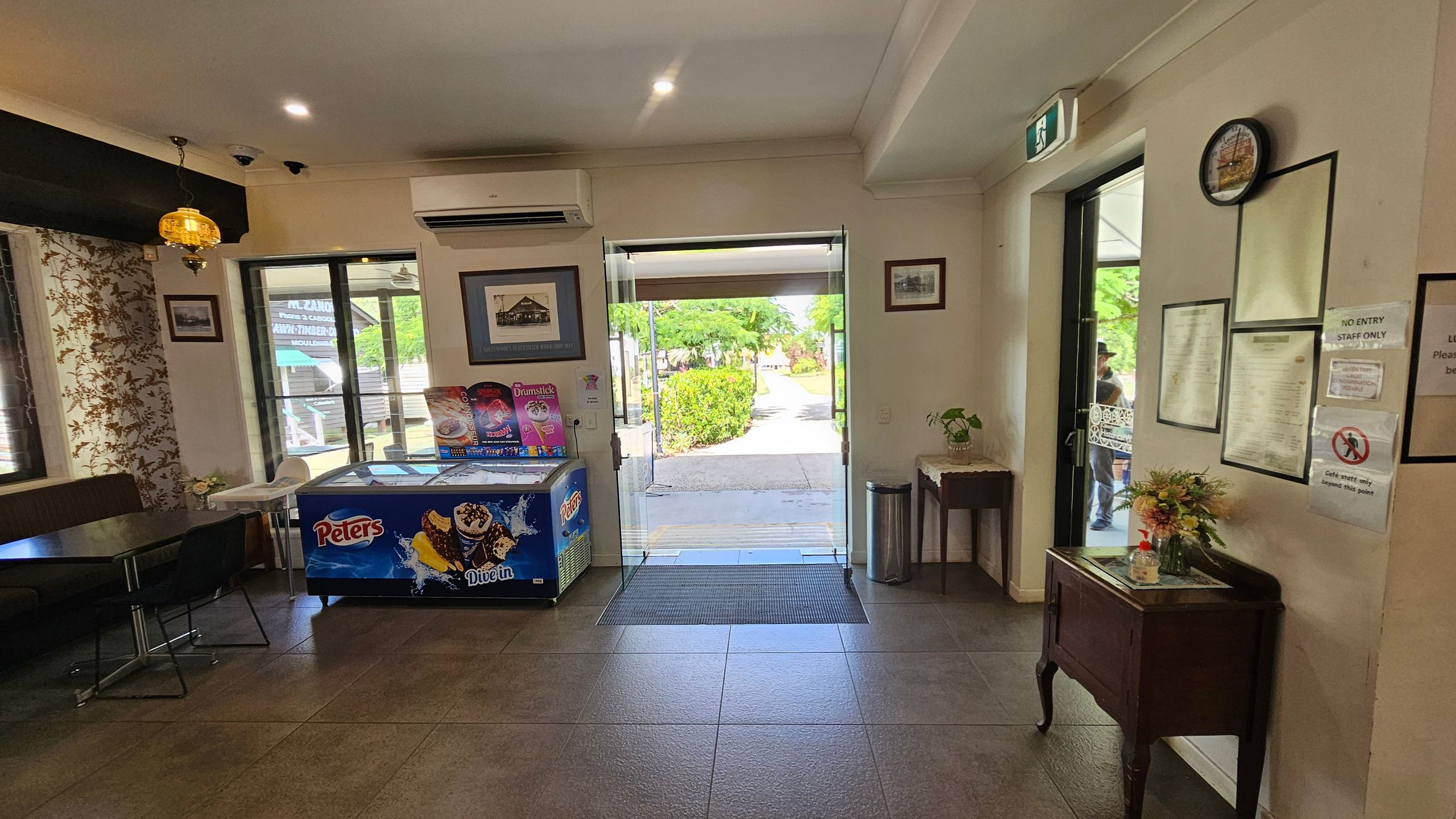 Inside a room with a tiled floor, a glass door in the centre opens to a path lined with greenery outside. On the left, a table with chairs and a decorative wall with floral patterns. An ice cream freezer with colourful advertisements is nearby. On the right, a small table with a plant and a wall displaying framed pictures and notices. A clock is mounted above. The ceiling has recessed lighting and a hanging lamp.