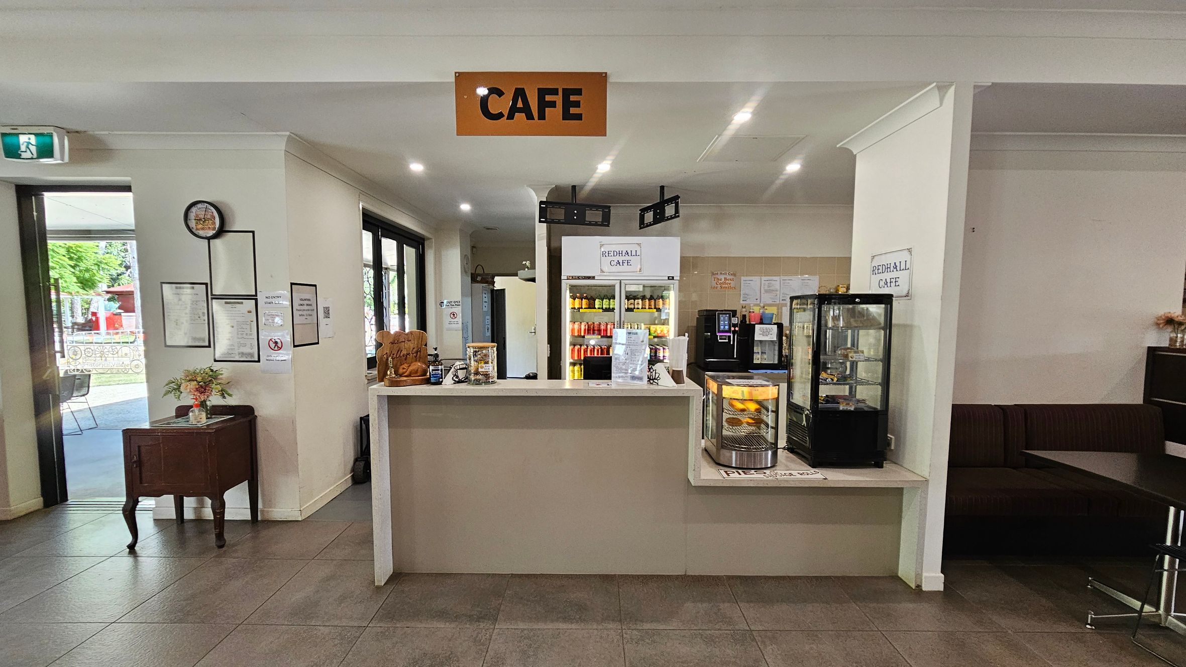 A café interior with a central counter displaying a small glass case containing pastries. Behind the counter, a fridge is stocked with various drinks. To the left, a small table with flowers and framed notices on the wall. An exit sign and glass doors lead outside. On the right, a seating area with a dark bench. The ceiling is white with recessed lighting, and the floor is tiled in a greyish tone. A sign above reads "CAFE".