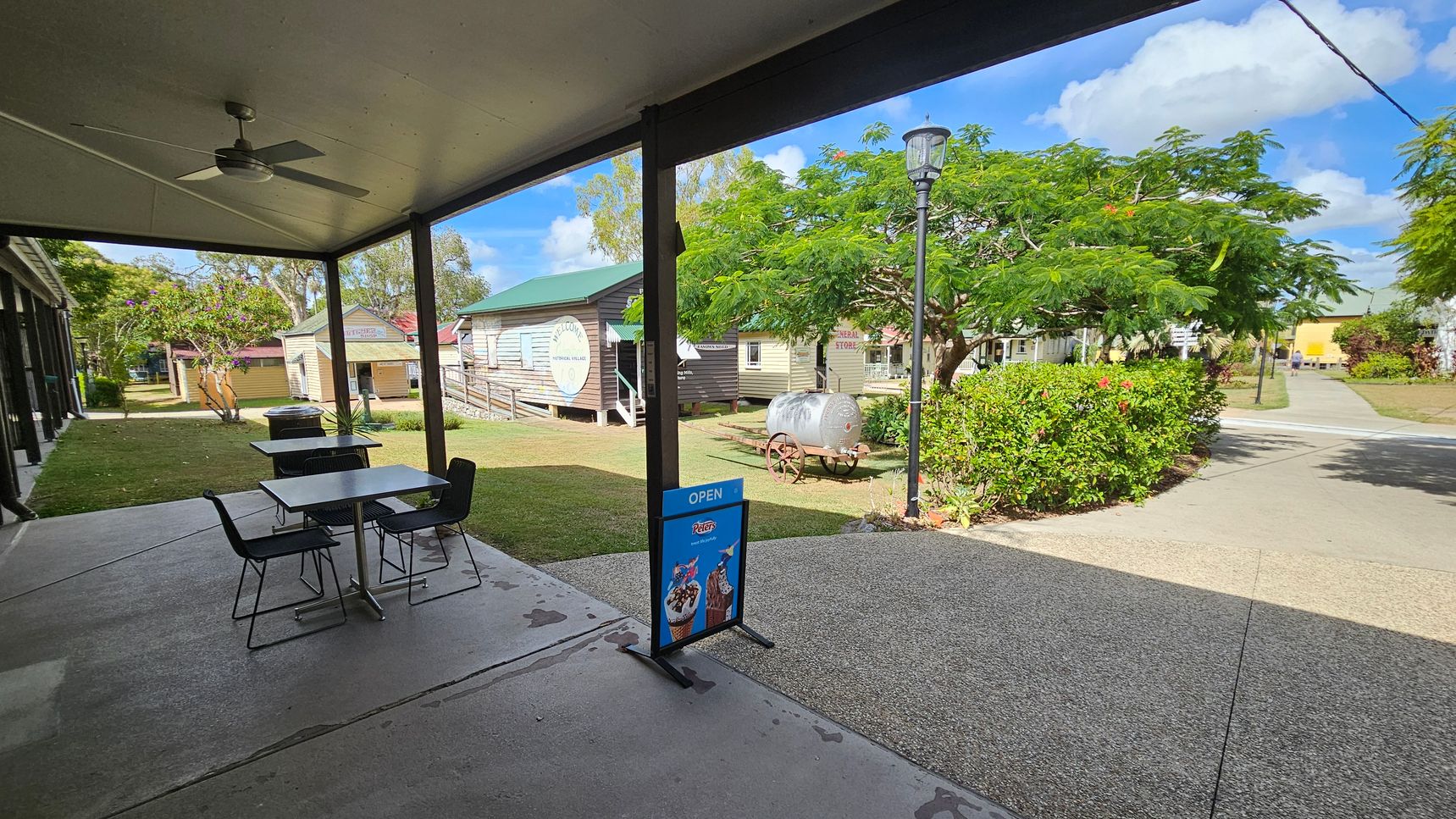 A covered outdoor area with a ceiling fan and two tables with chairs on a concrete floor. A sign with ice cream images and the word "OPEN" is in the foreground. In the background, there are wooden buildings with green roofs, one with a "Welcome" sign. A vintage water cart is near a tree with lush green foliage. A lamppost stands beside a path leading through a garden with bushes and flowers.