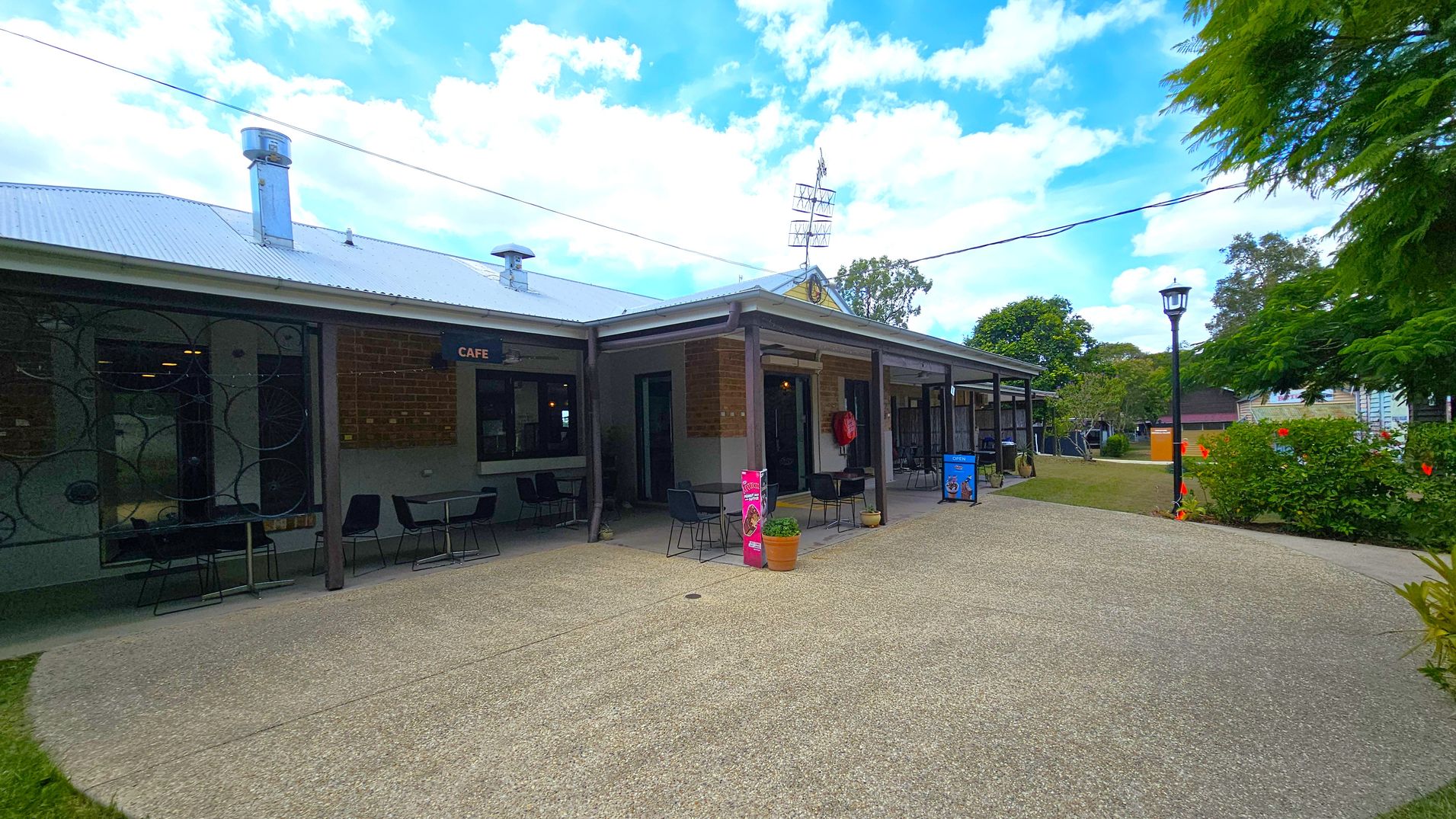 A single-storey building with a metal roof and several chimneys. The left side features a café with outdoor seating, including tables and chairs. The right side has a covered walkway with more seating and a fire extinguisher mounted on the wall. In the foreground, a paved area leads to a grassy section with a lamppost and bushes. Trees and additional buildings are visible in the background.