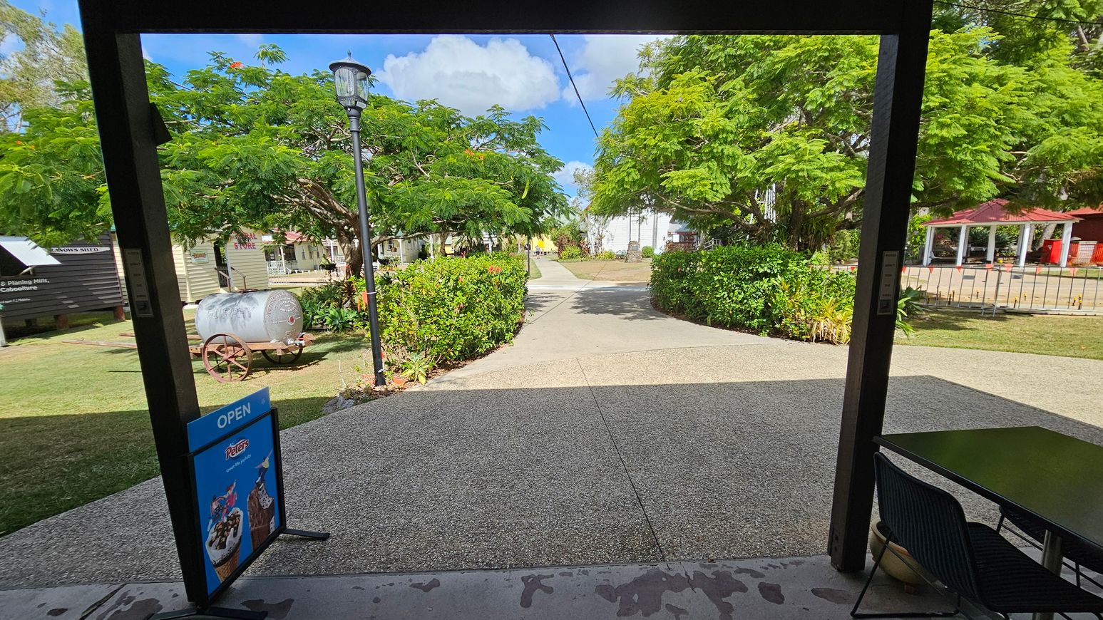 A view from a shaded area with a sign reading "OPEN" featuring ice cream images on the left. A table and chair are on the right. Beyond, a path leads through a garden with lush green trees and bushes. A vintage metal cart is on the left, and a lamppost stands nearby. In the background, there are small buildings and a red-roofed structure. The sky is partly cloudy.