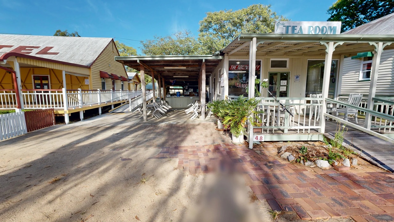 A quaint outdoor setting with a tea room on the right, featuring a porch with white railings and potted plants. The entrance has a sign above it. In the centre, there is a covered seating area with white chairs and tables. On the left, a building with a sign partially visible, possibly reading "HOTEL," has a wooden exterior and a ramp leading up to it. The ground is a mix of sand and brick paving, and trees are visible in the background.